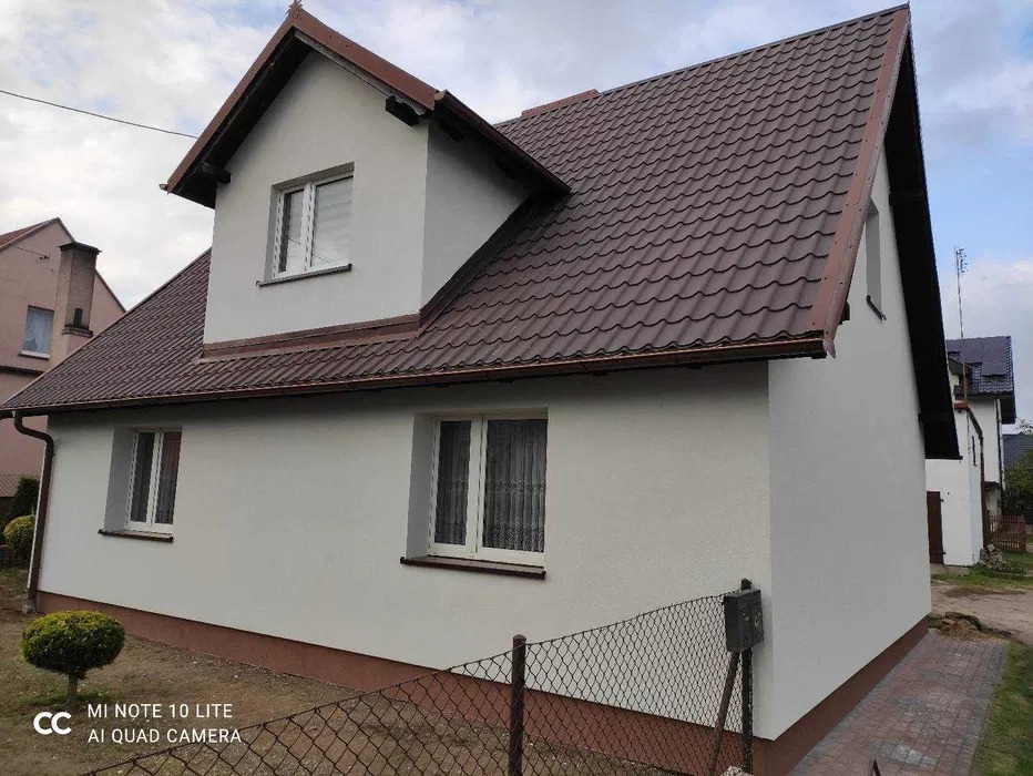 Jasna elewacja domu jednorodzinnego z brązowym dachem i podmurówką, widok z boku, ogrodzenie z siatki z przodu, krzew ozdobny po lewej.