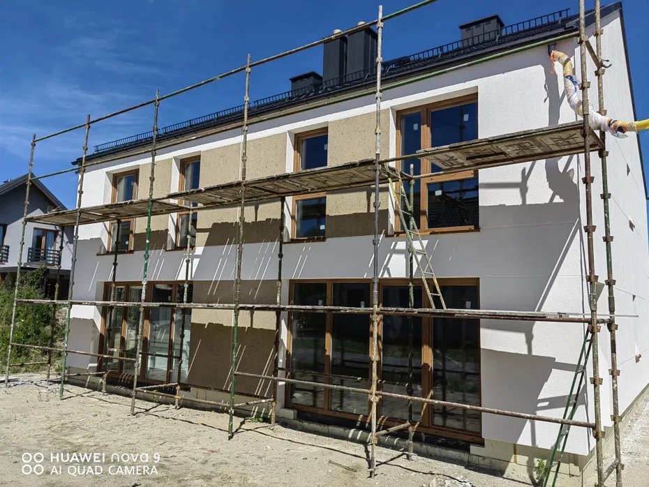 Nowy dom w trakcie ocieplania, z widocznymi rusztowaniami i fragmentem elewacji pokrytej tynkiem strukturalnym w odcieniach bieli i beżu, na tle błękitnego nieba.