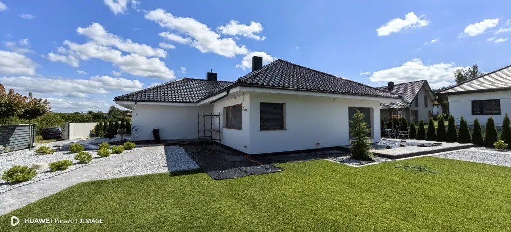 Biała elewacja domu jednorodzinnego z ciemną dachówką, widok z boku, ogród z trawnikiem i ścieżką z kamienia. W tle błękitne niebo z chmurami.