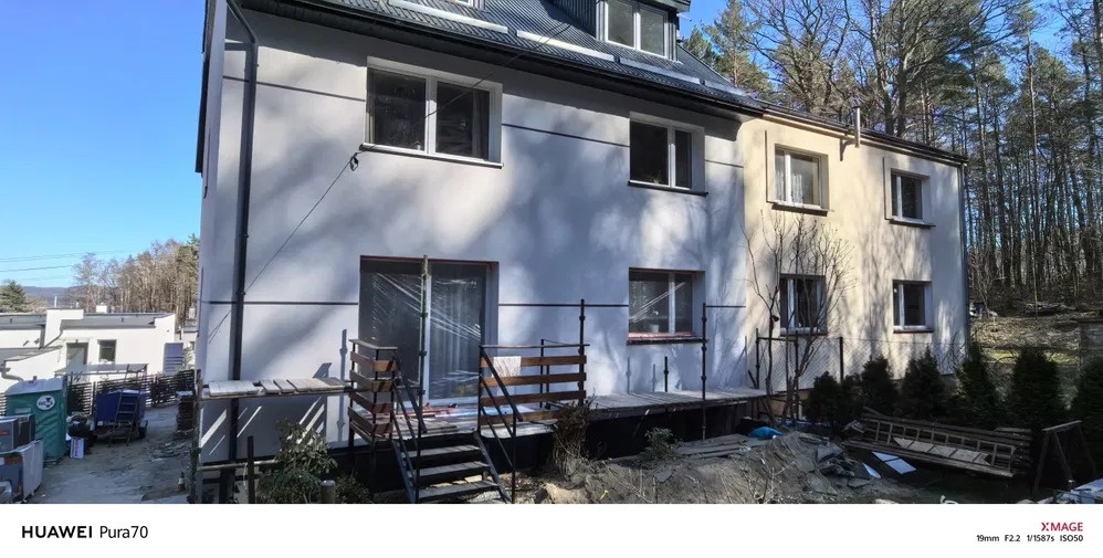 Dwukondygnacyjny dom w trakcie tynkowania elewacji, z widocznymi rusztowaniami i świeżo otynkowaną fasadą w odcieniach bieli i beżu, w słoneczny dzień.