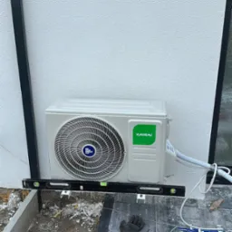 Biała zewnętrzna jednostka klimatyzacji Kaisai zamontowana na ścianie budynku, wypoziomowana poziomicą. Na ziemi narzędzia i robocze rękawice.