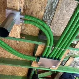 Zielone, elastyczne rury wentylacyjne przymocowane do drewnianej konstrukcji dachu, widoczne elementy montażowe i fragmenty izolacji. Ujęcie z dołu, z perspektywy instalatora.