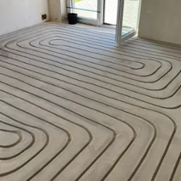 Ułożone rury ogrzewania podłogowego w surowym pomieszczeniu z otwartym oknem balkonowym i widokiem na ogród. Widoczne złącza na ścianie.