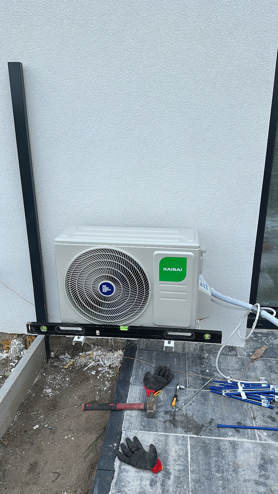 Biała zewnętrzna jednostka klimatyzacji Kaisai zamontowana na ścianie budynku, wypoziomowana poziomicą. Na ziemi narzędzia i robocze rękawice.