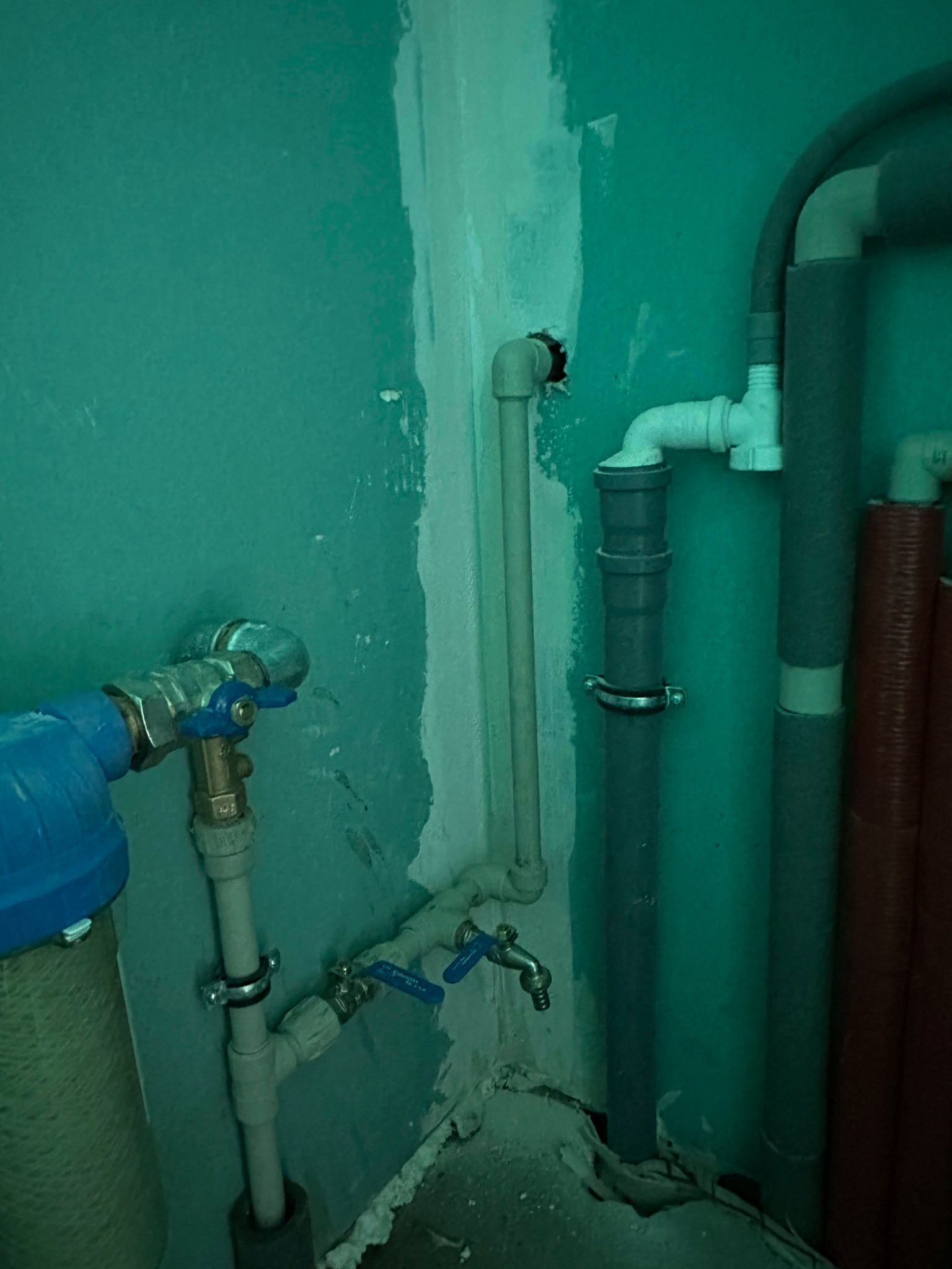 Fragment instalacji hydraulicznej w pomieszczeniu z turkusowymi ścianami. Widoczne rury z zaworami, filtr wody i elementy mocujące. Ujęcie z bliska, brak ostrości.