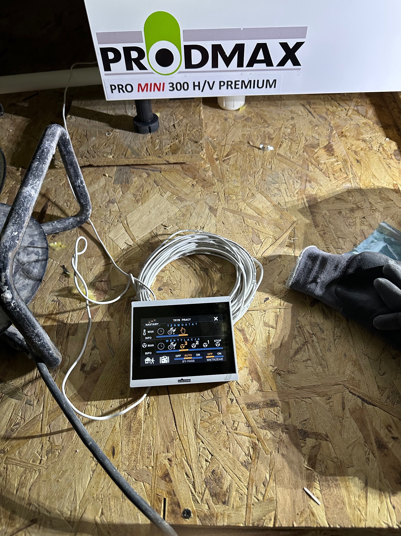 Panel sterowania Prodmax Pro Mini 300 H/V Premium, z widocznymi kablami i rękawicami roboczymi, na tle płyty OSB. Kontroler instalacji grzewczej w trakcie podłączania.