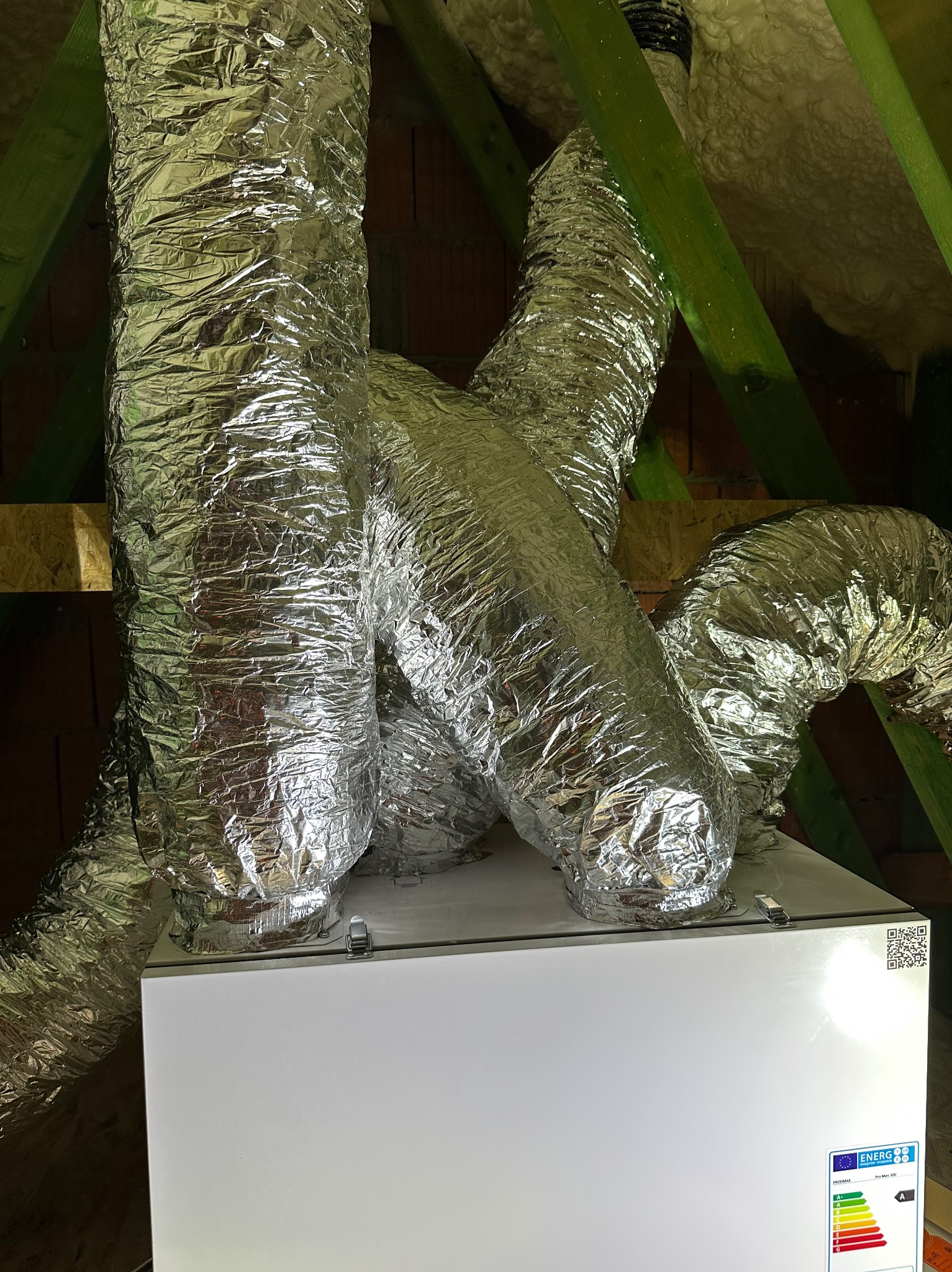 Biała centrala wentylacyjna z podłączonymi, izolowanymi przewodami wentylacyjnymi w folii aluminiowej, zamontowana na poddaszu z widoczną więźbą dachową.