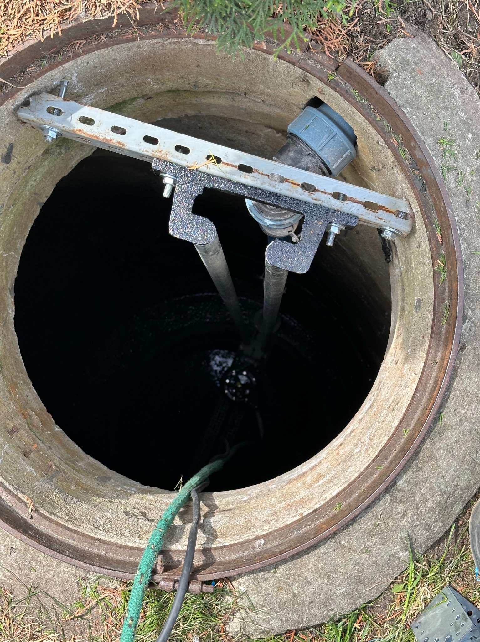 Otwarty betonowy właz studzienki kanalizacyjnej z zamontowanym metalowym uchwytem i rurami wewnątrz. Widoczne przewody. Ujęcie z góry.