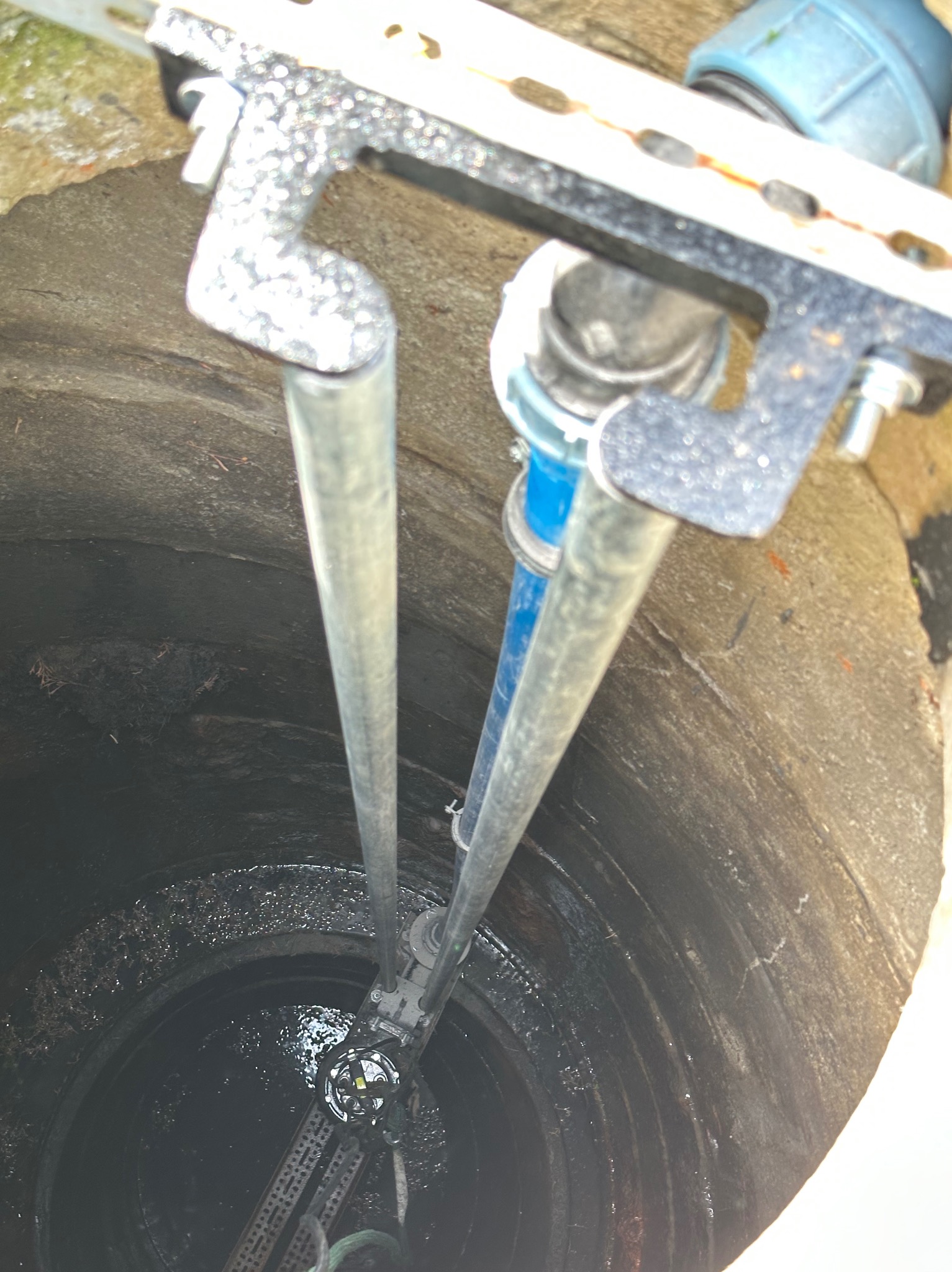 Wnętrze studni z zanurzonymi rurami i elementami instalacji. Widok z góry, oświetlenie naturalne. Metalowe i plastikowe elementy instalacji w surowym otoczeniu studni.