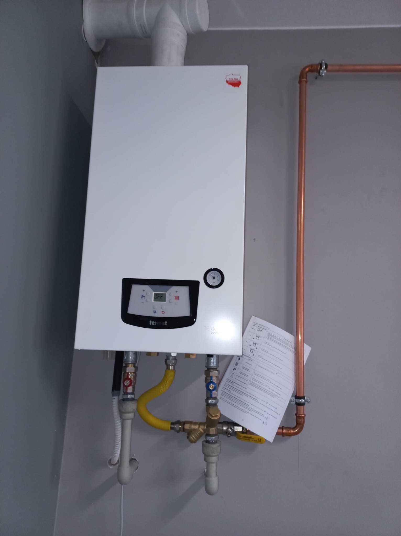 Biały piec gazowy Termet z miedzianą instalacją na szarej ścianie. Widoczne podłączenia, rury odprowadzające spaliny i dokumentacja techniczna.