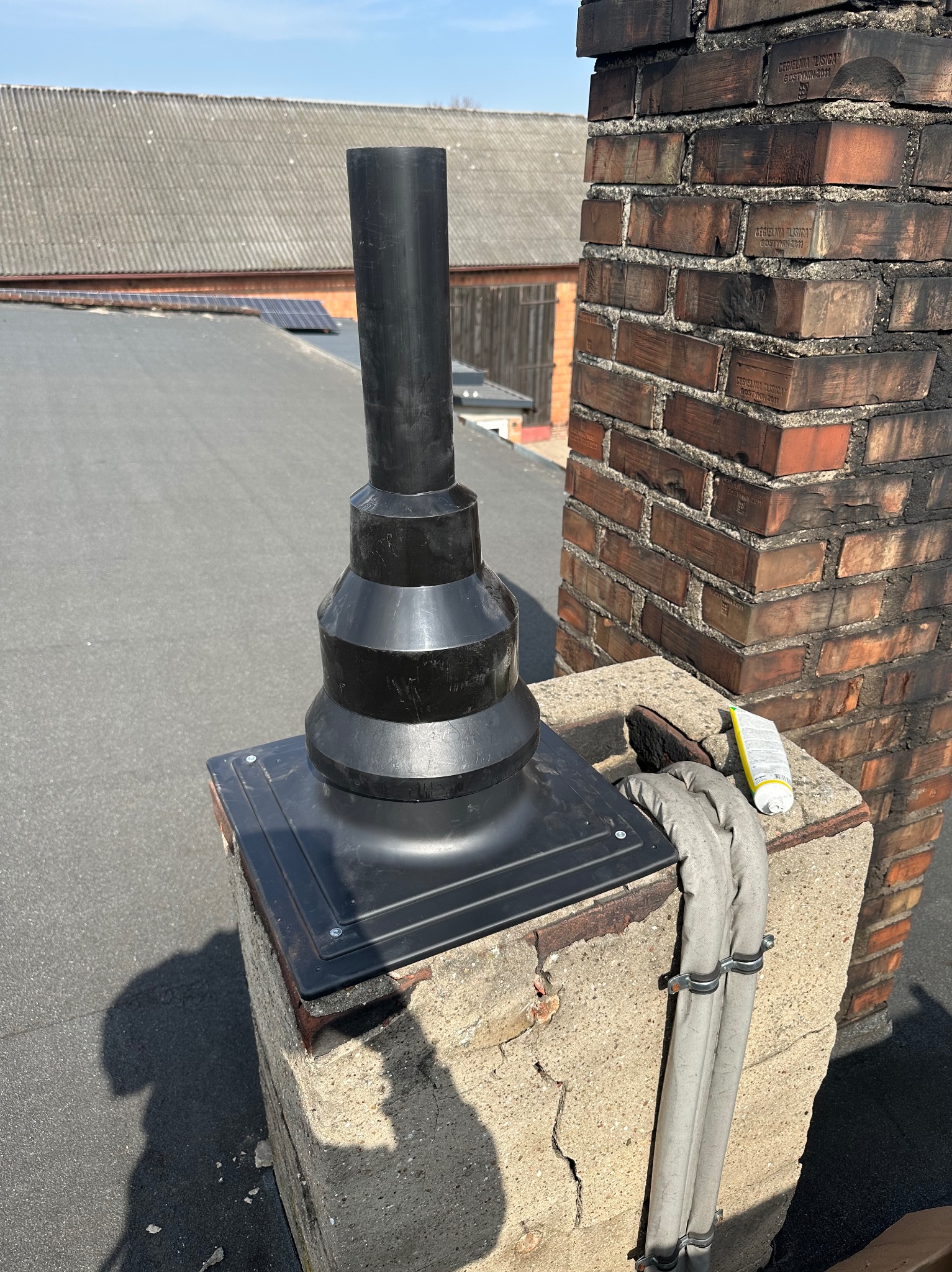 Czarny komin wentylacyjny na betonowej podstawie, obok ceglany komin. Na dachu widoczne panele słoneczne i budynki gospodarcze. Słoneczny dzień.