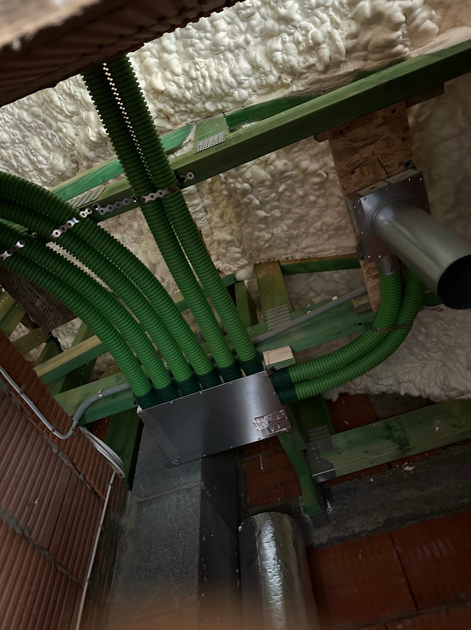 Zielone rury wentylacyjne w izolowanym poddaszu, połączone z metalową skrzynką. Fragment drewnianej konstrukcji dachu. Widok instalacji od dołu, z bliska.