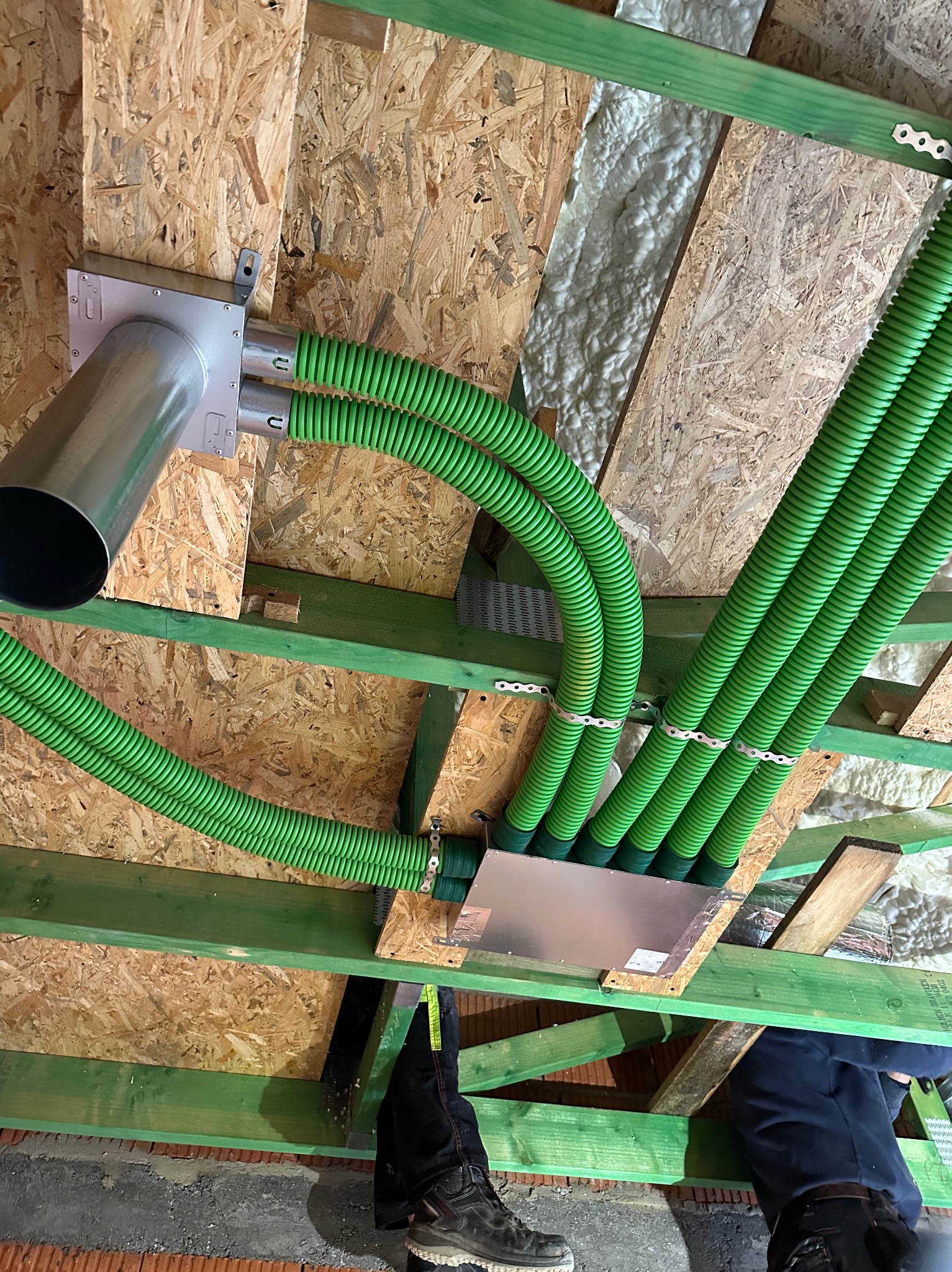 Zielone, elastyczne rury wentylacyjne przymocowane do drewnianej konstrukcji dachu, widoczne elementy montażowe i fragmenty izolacji. Ujęcie z dołu, z perspektywy instalatora.