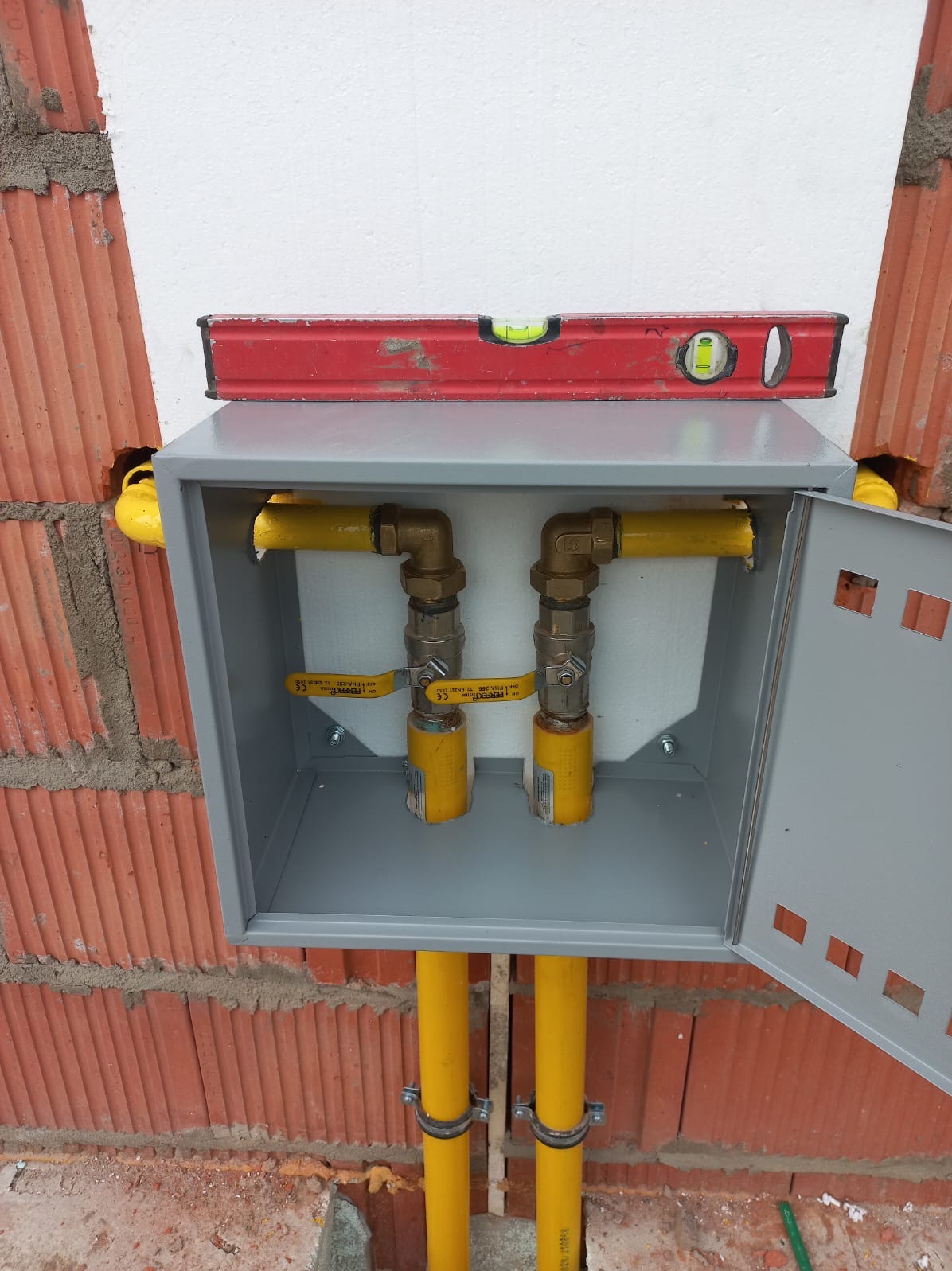 Szara skrzynka z zaworami gazowymi i żółtymi rurami, zamontowana na ceglanej ścianie. Na skrzynce leży poziomica.