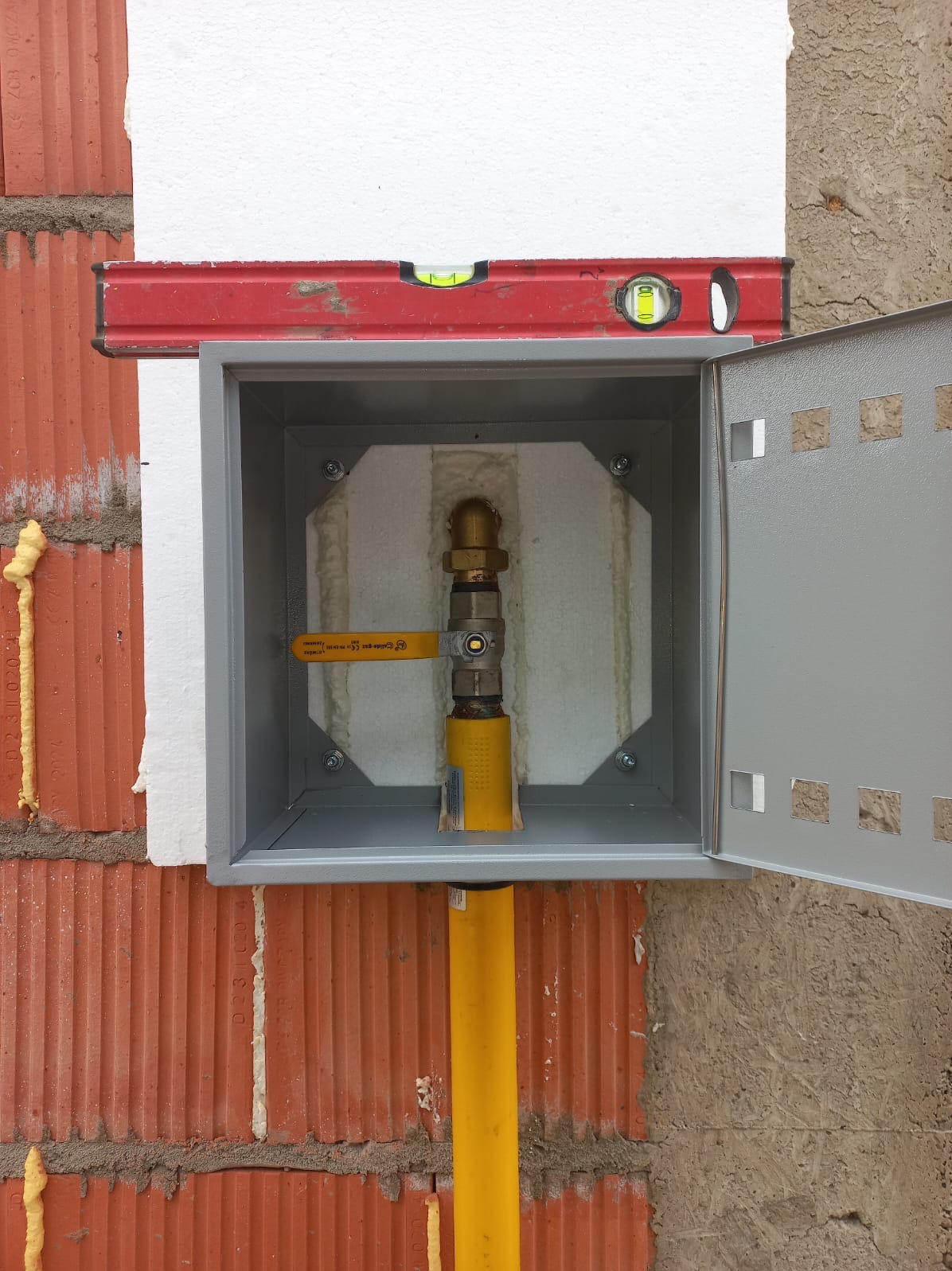Szara skrzynka gazowa z otwartymi drzwiczkami, zamontowana na ścianie z cegły. Widoczny żółty zawór i rura gazowa. Na górze skrzynki poziomica.