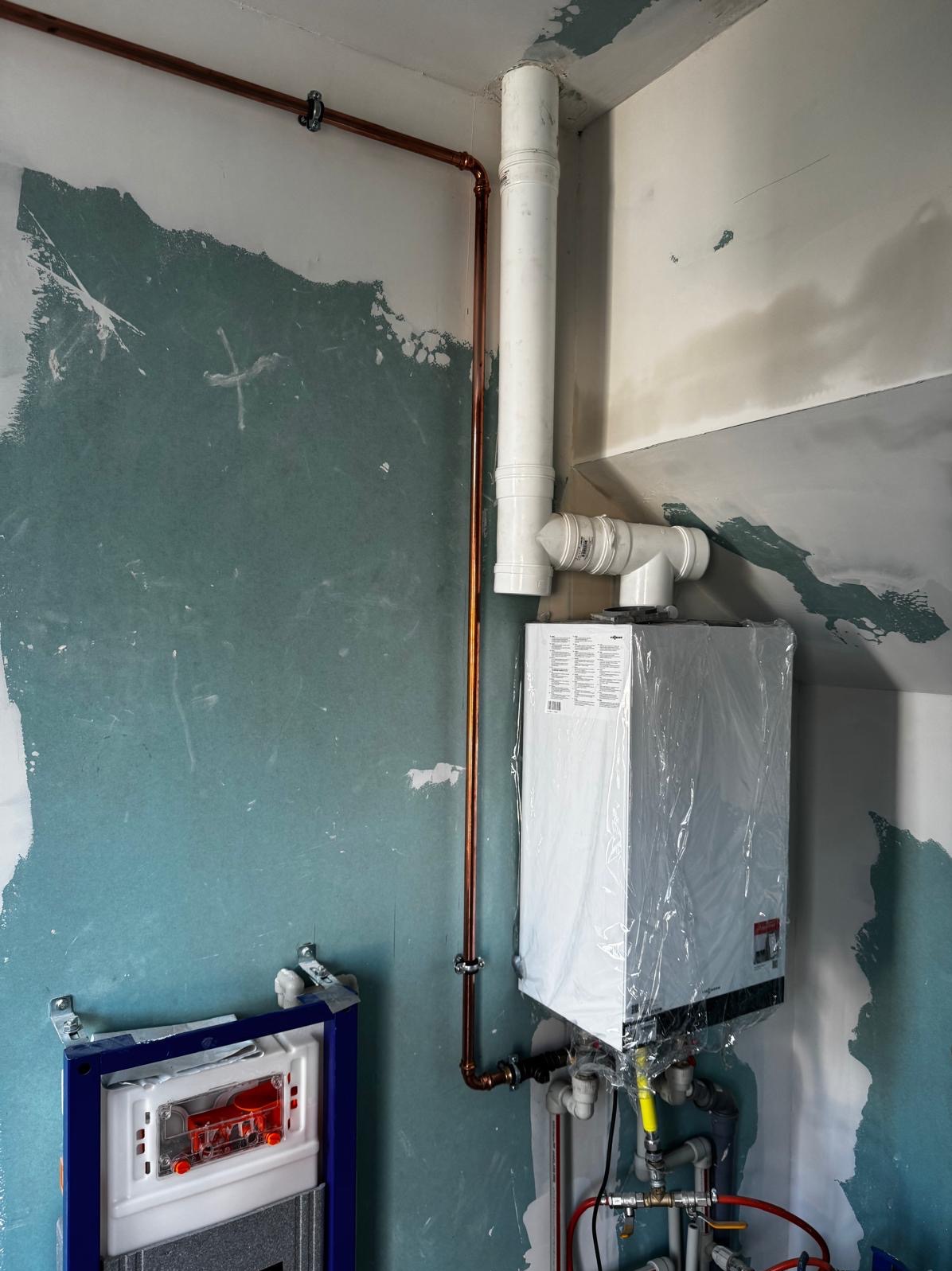 Nowy piec gazowy Viessmann w folii ochronnej, podłączony do miedzianej instalacji, z widocznymi rurami odprowadzającymi spaliny. Ściana w trakcie wykończenia.