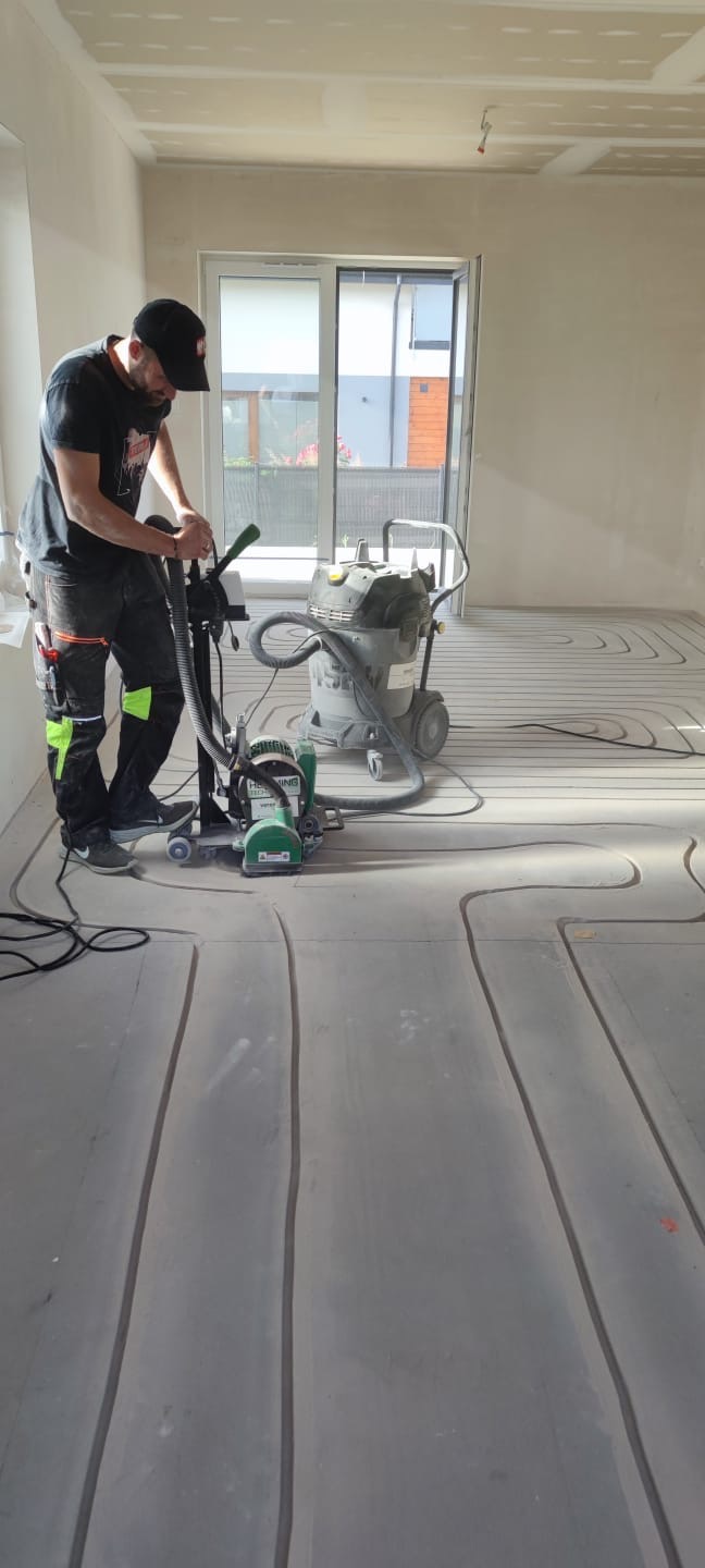 Frezowanie podłoża pod ogrzewanie podłogowe: mężczyzna w czapce frezuje podłogę specjalistycznym sprzętem, widoczne rowki pod rury.