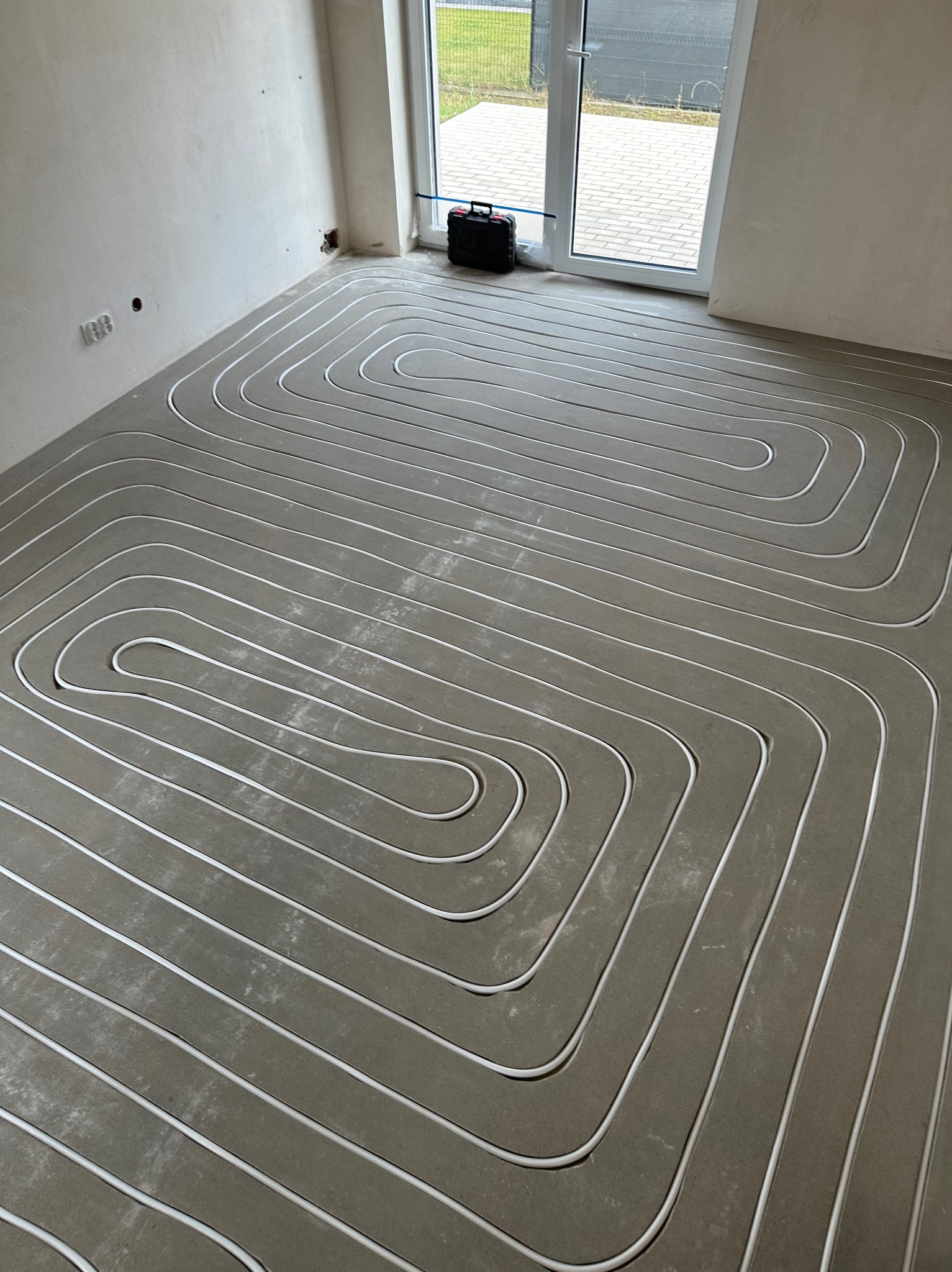 Białe rury ogrzewania podłogowego ułożone w pętle na szarym podkładzie w surowym wnętrzu. Widoczne gniazdka elektryczne i okno tarasowe. Czarna skrzynka narzędziowa w rogu.