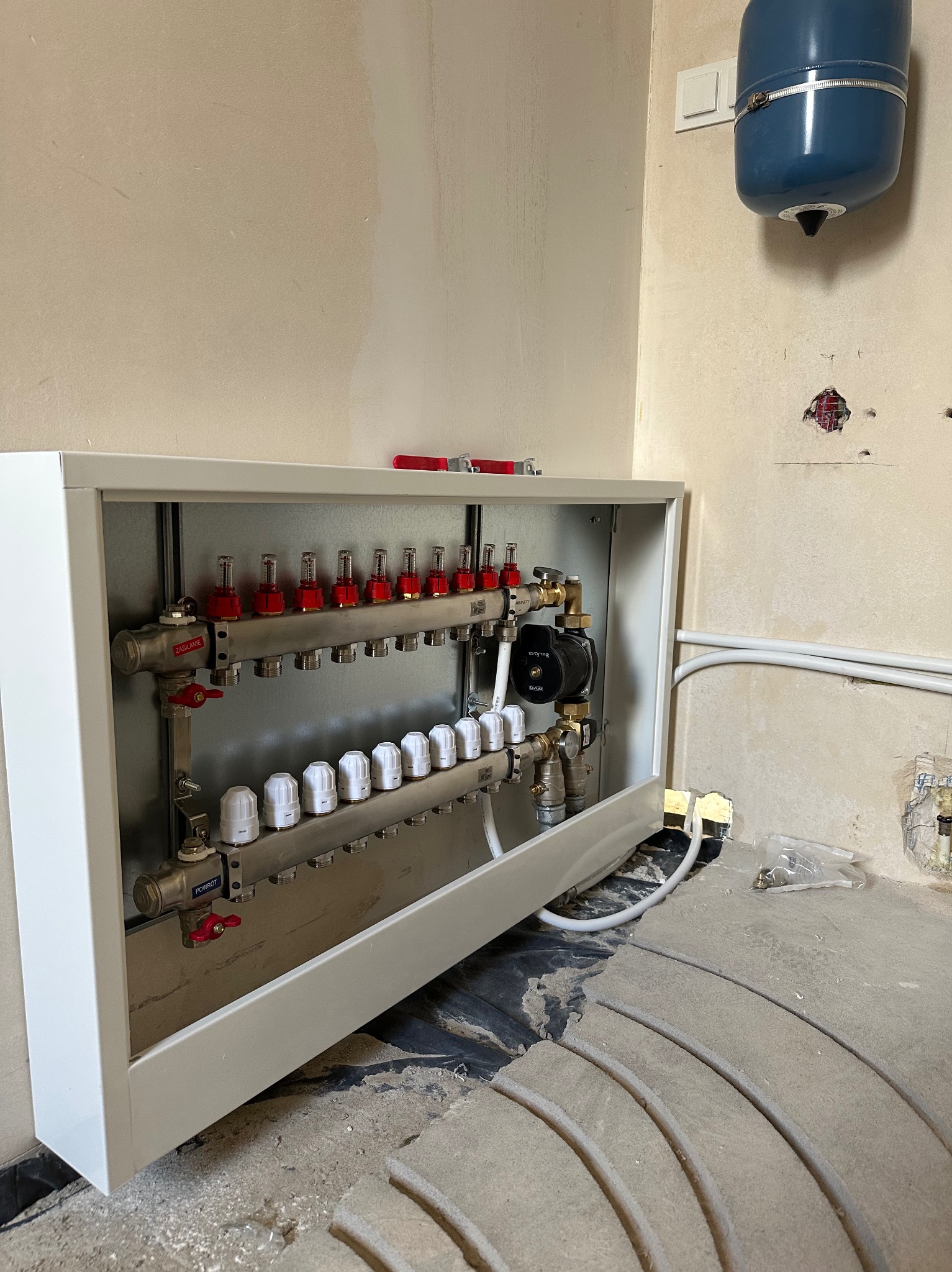 Rozdzielacz ogrzewania podłogowego w obudowie, widoczne rury i elementy instalacji. Na podłodze fragmenty izolacji termicznej. Ściany w stanie surowym.
