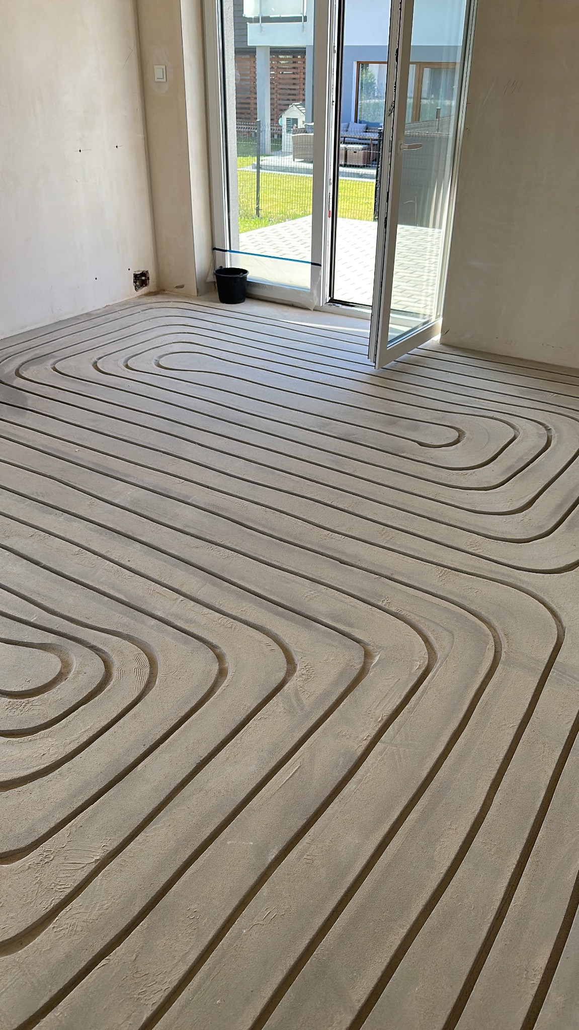 Ułożone rury ogrzewania podłogowego w surowym pomieszczeniu z otwartym oknem balkonowym i widokiem na ogród. Widoczne złącza na ścianie.