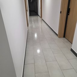Lerion Cleaning - Wnętrze korytarza z jasnymi płytkami podłogowymi i białymi ścianami. Widoczne drzwi z numerami mieszkań. Minimalistyczny design, czystość i porządek.