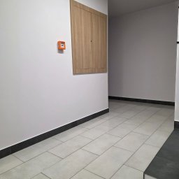 Lerion Cleaning - Wnętrze korytarza po remoncie: jasne płytki podłogowe, białe ściany z brązową szafką i pomarańczowym czujnikiem dymu. Czyste, minimalistyczne wykończenie.