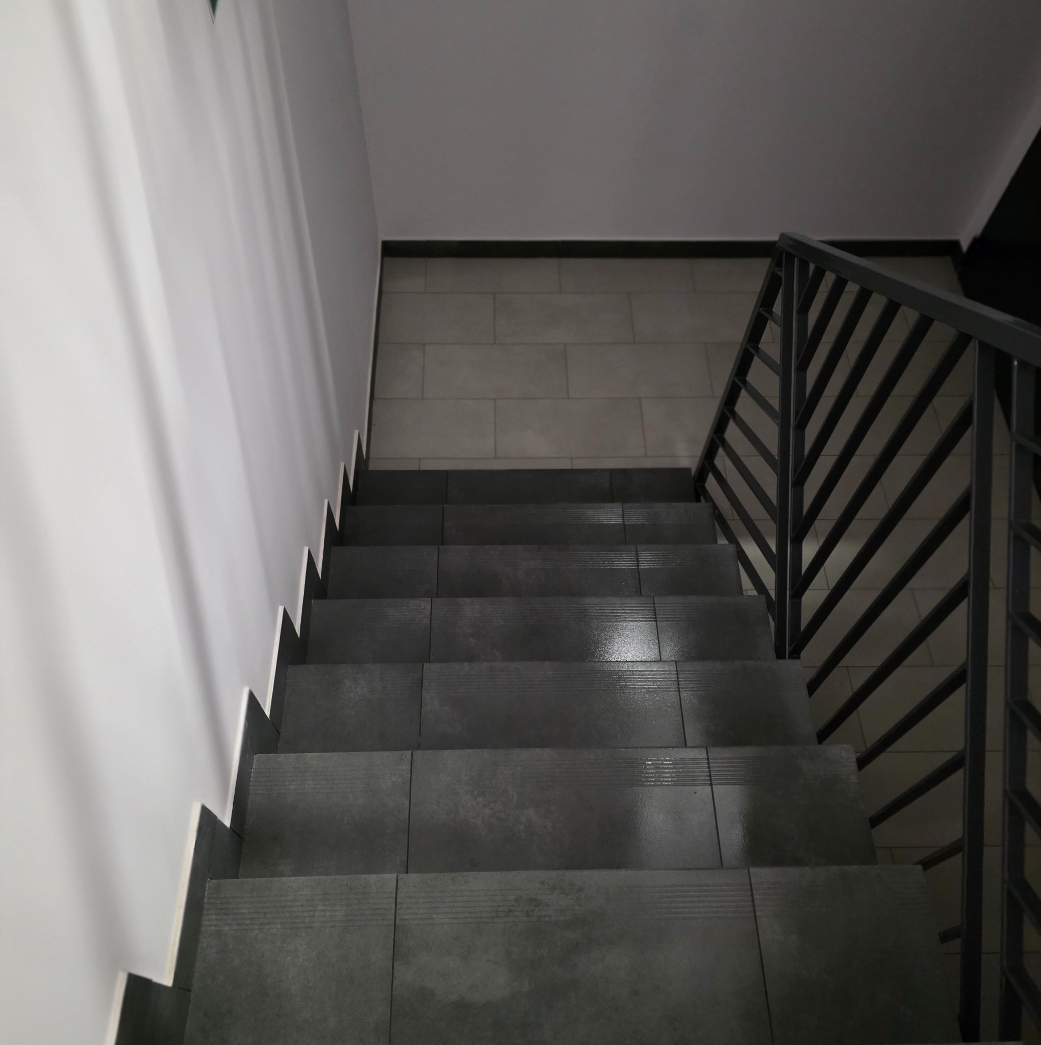 Schody wyłożone szarymi płytkami z metalową barierką, widok z góry. Świeżo umyte, mokre płytki schodowe z delikatnym połyskiem. Minimalistyczny design.