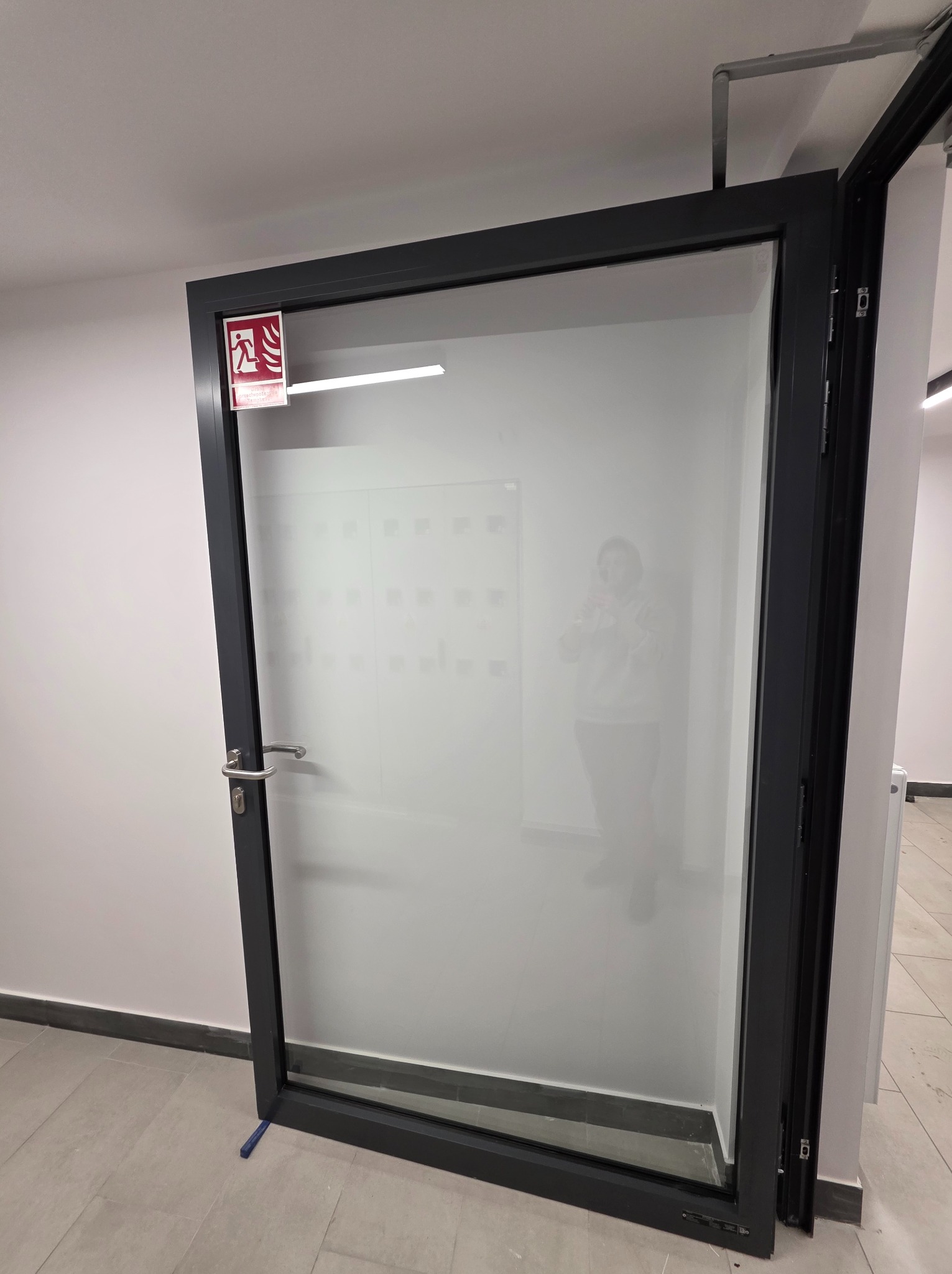 Szklane drzwi ewakuacyjne z czarną ramą, widoczne z boku, z naklejką informacyjną w górnym rogu. Na podłodze płytki, ściany białe. Widoczne odbicie osoby w szybie.