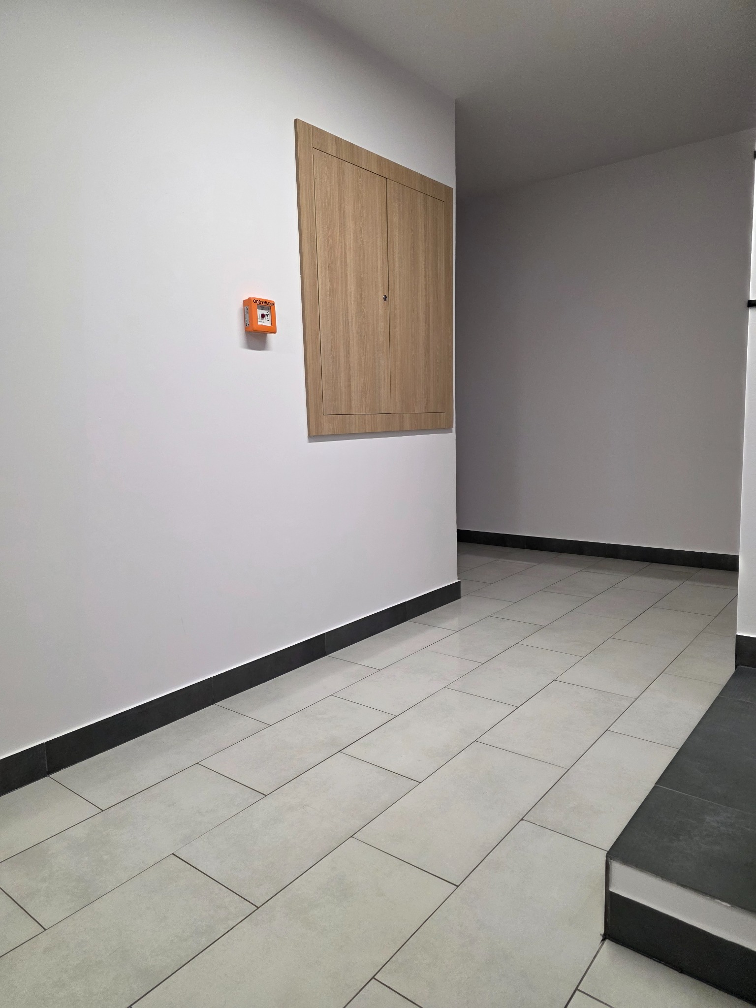 Wnętrze korytarza po remoncie: jasne płytki podłogowe, białe ściany z brązową szafką i pomarańczowym czujnikiem dymu. Czyste, minimalistyczne wykończenie.