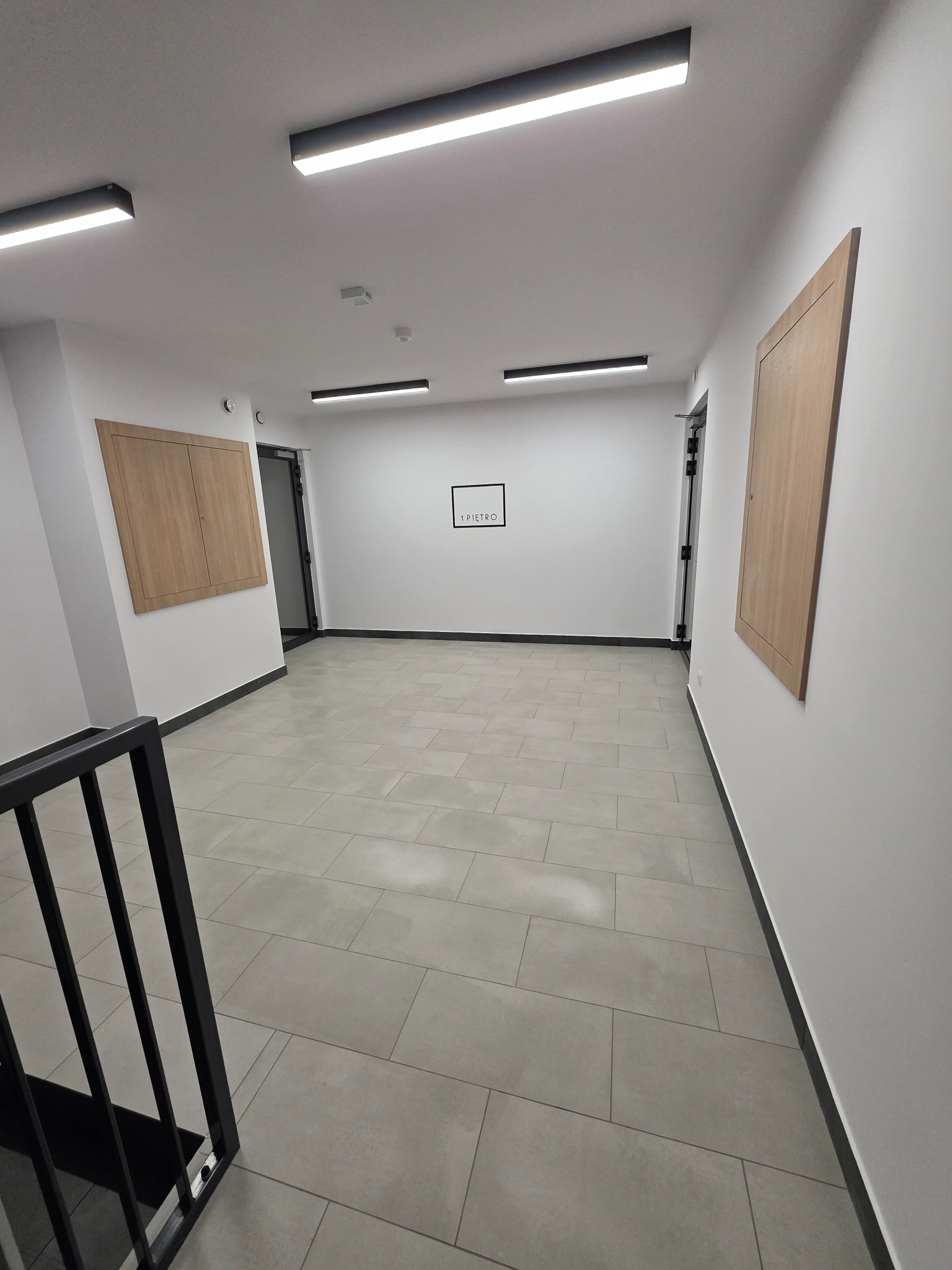 Wnętrze korytarza z szarymi płytkami podłogowymi i białymi ścianami. Widoczne drzwi, oświetlenie sufitowe i fragment schodów z czarną barierką. Minimalistyczny design.