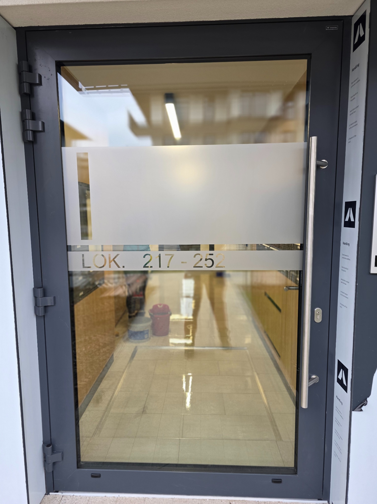 Szklane drzwi z numerem lokalu 2.17-252, widok na wnętrze z wiadrem i mopem. Świeżo umyte, lśniące szkło. Widoczne odbicia światła.