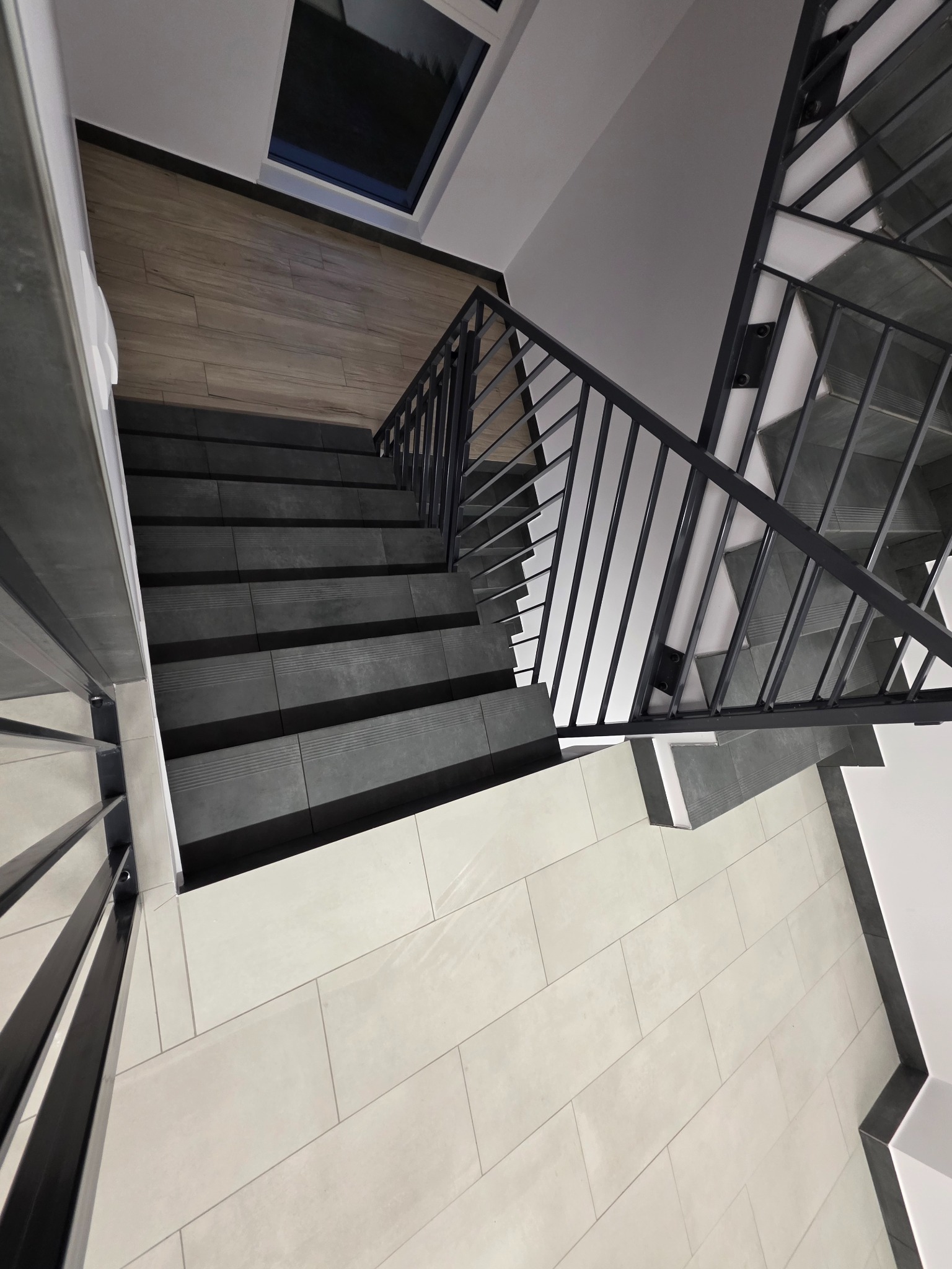 Nowoczesne schody z ciemnoszarymi stopniami i metalową balustradą, widok z góry na jasną podłogę. Minimalistyczny design z oknem w tle.