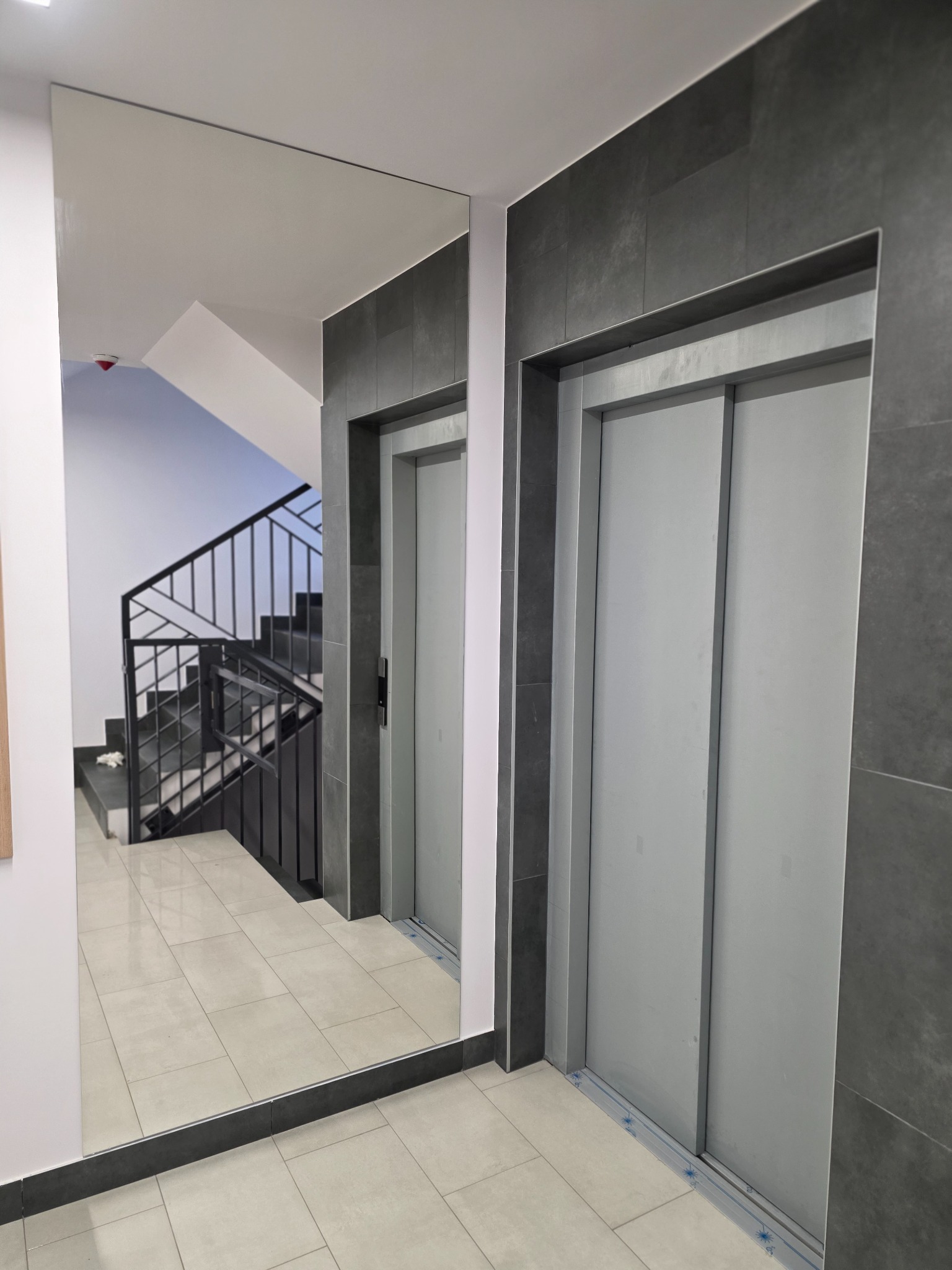 Wnętrze klatki schodowej z lustrem, w którym odbija się schody. Obok widoczne dwie windy z szarymi drzwiami i ciemnoszare płytki na ścianach.