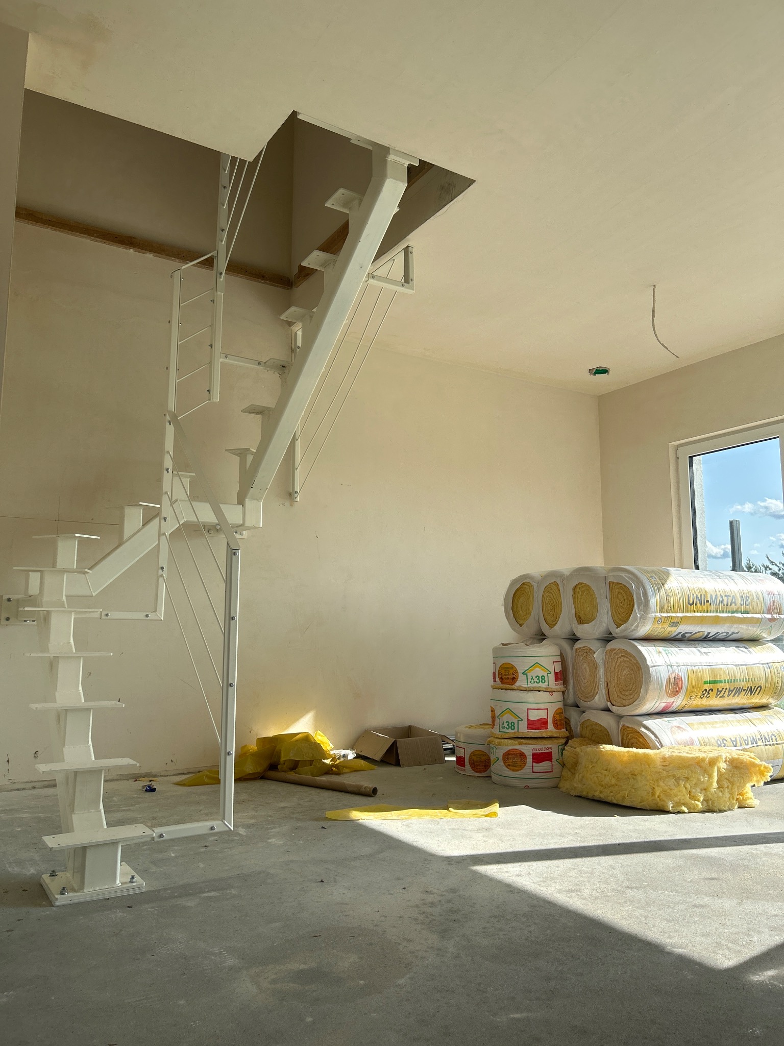 Wnętrze w trakcie prac wykończeniowych: białe schody metalowe, ocieplenie w rolkach, surowe ściany i podłoga. Okno z widokiem na zewnątrz.