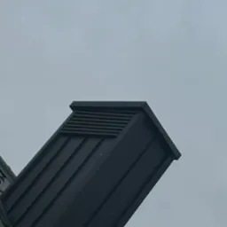 Nowoczesny, czarny kominek wentylacyjny na dachu z blachy na tle pochmurnego nieba. Widok z dołu, podkreślający minimalistyczny design.