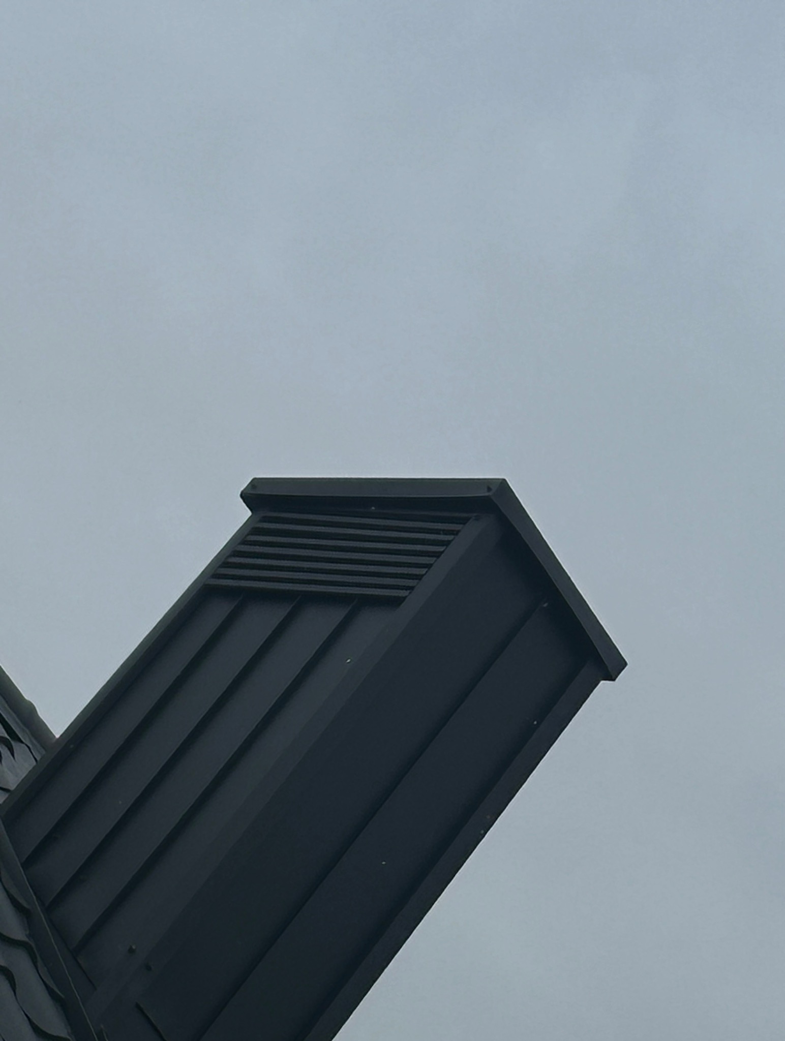 Nowoczesny, czarny kominek wentylacyjny na dachu z blachy na tle pochmurnego nieba. Widok z dołu, podkreślający minimalistyczny design.