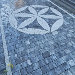 Ułożona kostka brukowa w szaro-białej kolorystyce z dekoracyjnym motywem kwiatowym na podjeździe. W tle budynek z elementami zieleni.