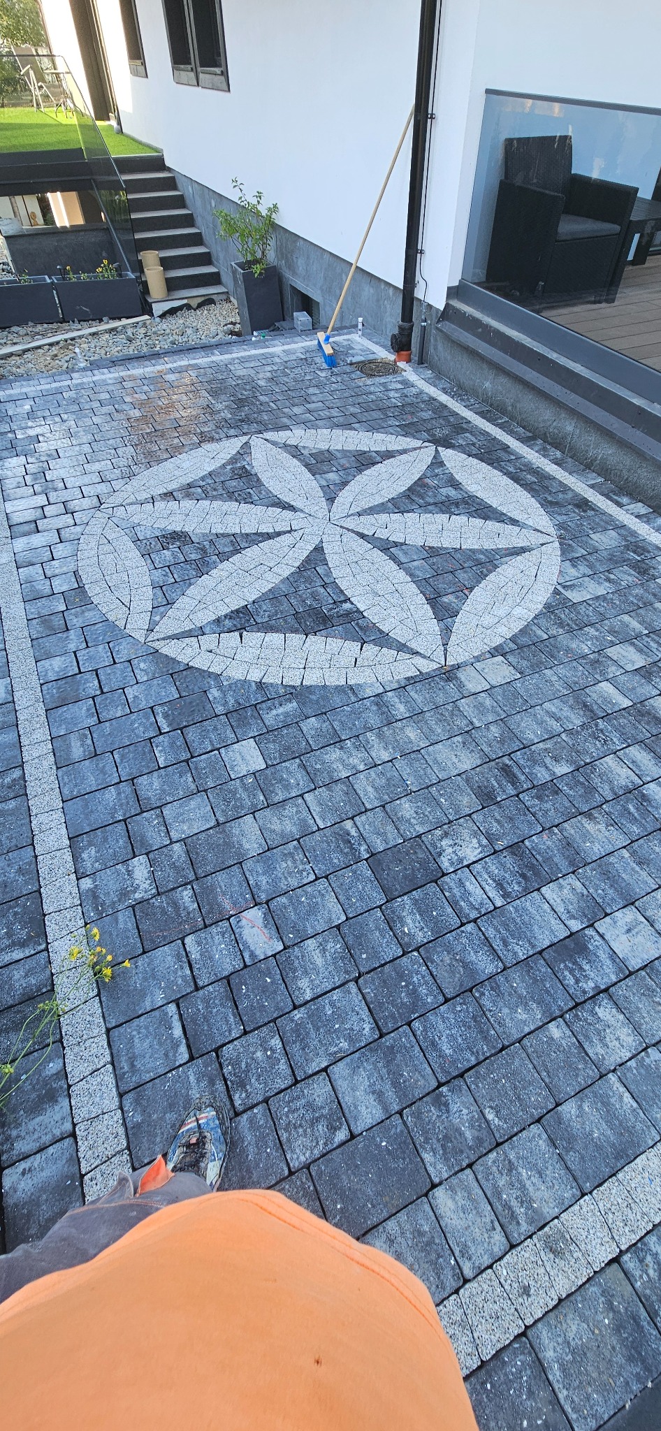 Ułożona kostka brukowa w szaro-białej kolorystyce z dekoracyjnym motywem kwiatowym na podjeździe. W tle budynek z elementami zieleni.