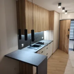 Nowoczesna kuchnia z drewnianymi szafkami wiszącymi, czarnym blatem i minimalistycznym oświetleniem. Widoczny fragment wyspy kuchennej i drewniana podłoga.