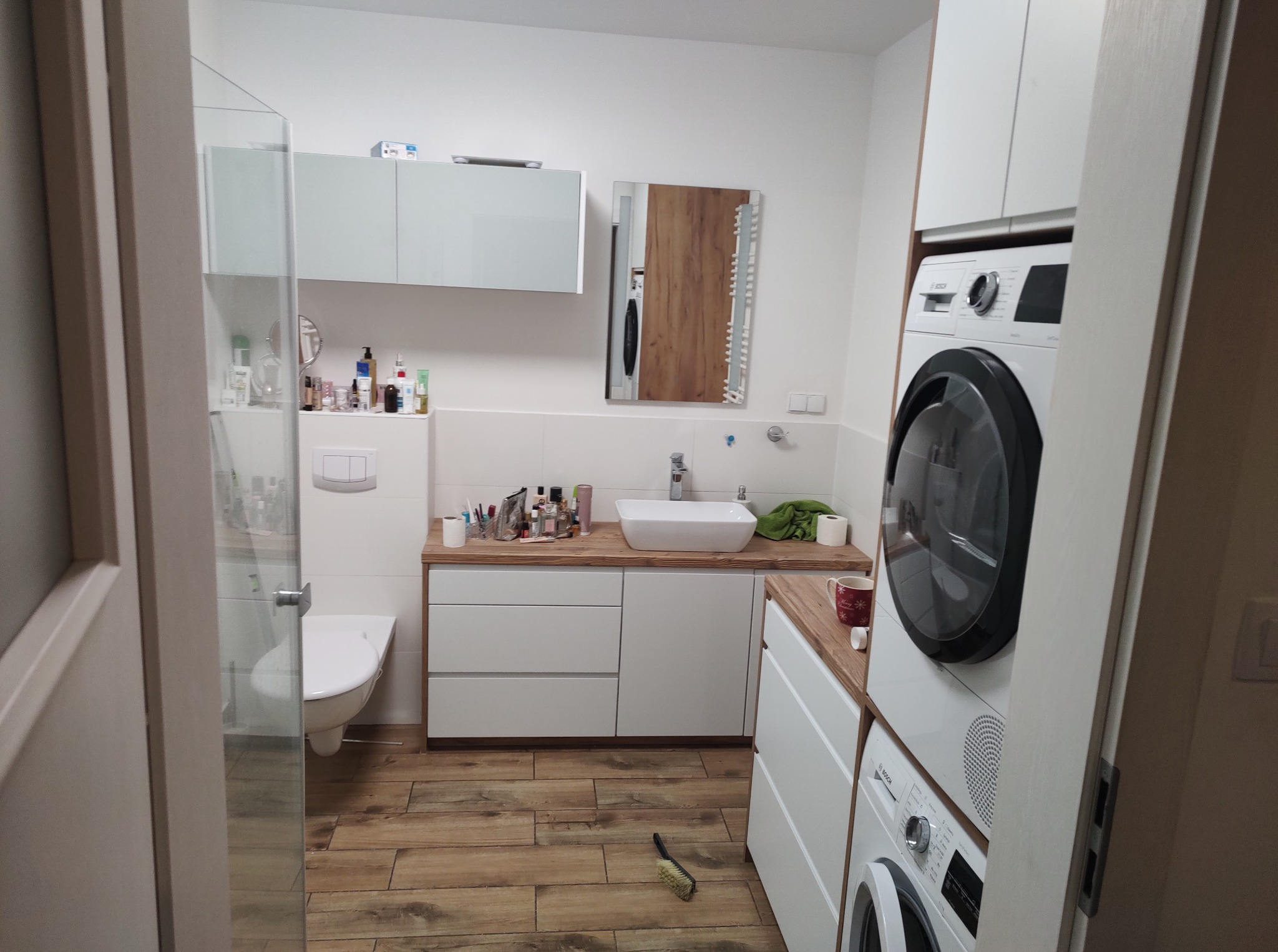 Nowoczesna łazienka z drewnianą podłogą i blatem, białymi szafkami, pralką i suszarką w zabudowie. Widok zza uchylonych drzwi.