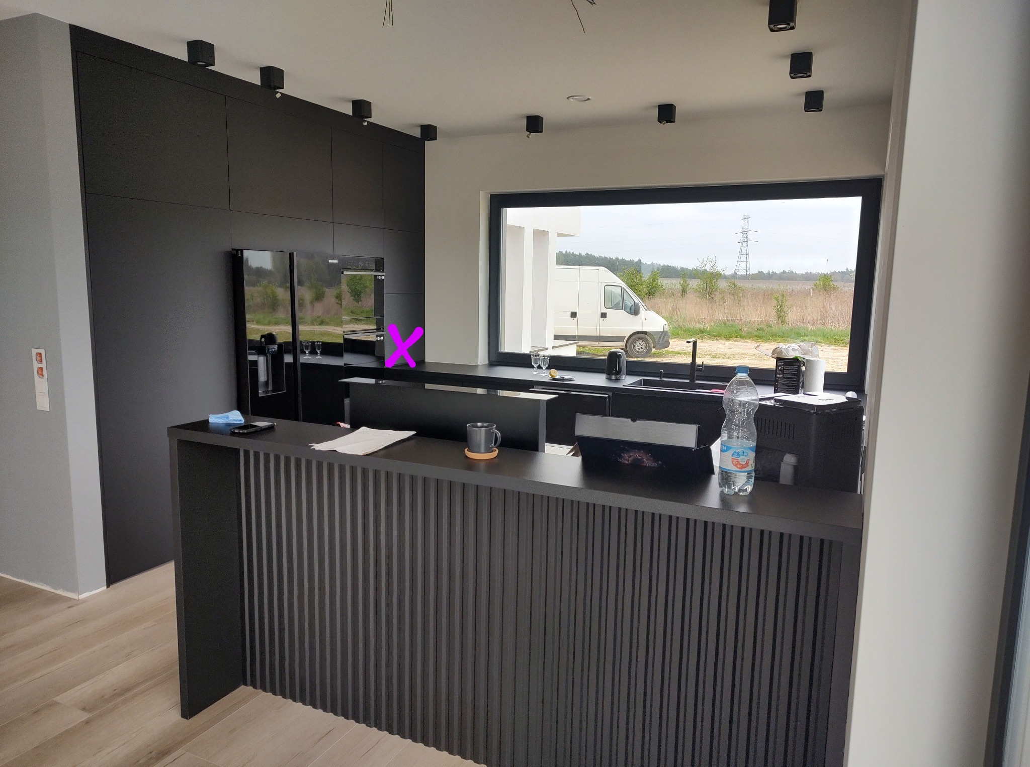 Nowoczesna, czarna zabudowa kuchenna z wyspą i oknem z widokiem na zewnątrz. Minimalistyczny design z pionowymi żłobieniami na wyspie. Oświetlenie punktowe na suficie.