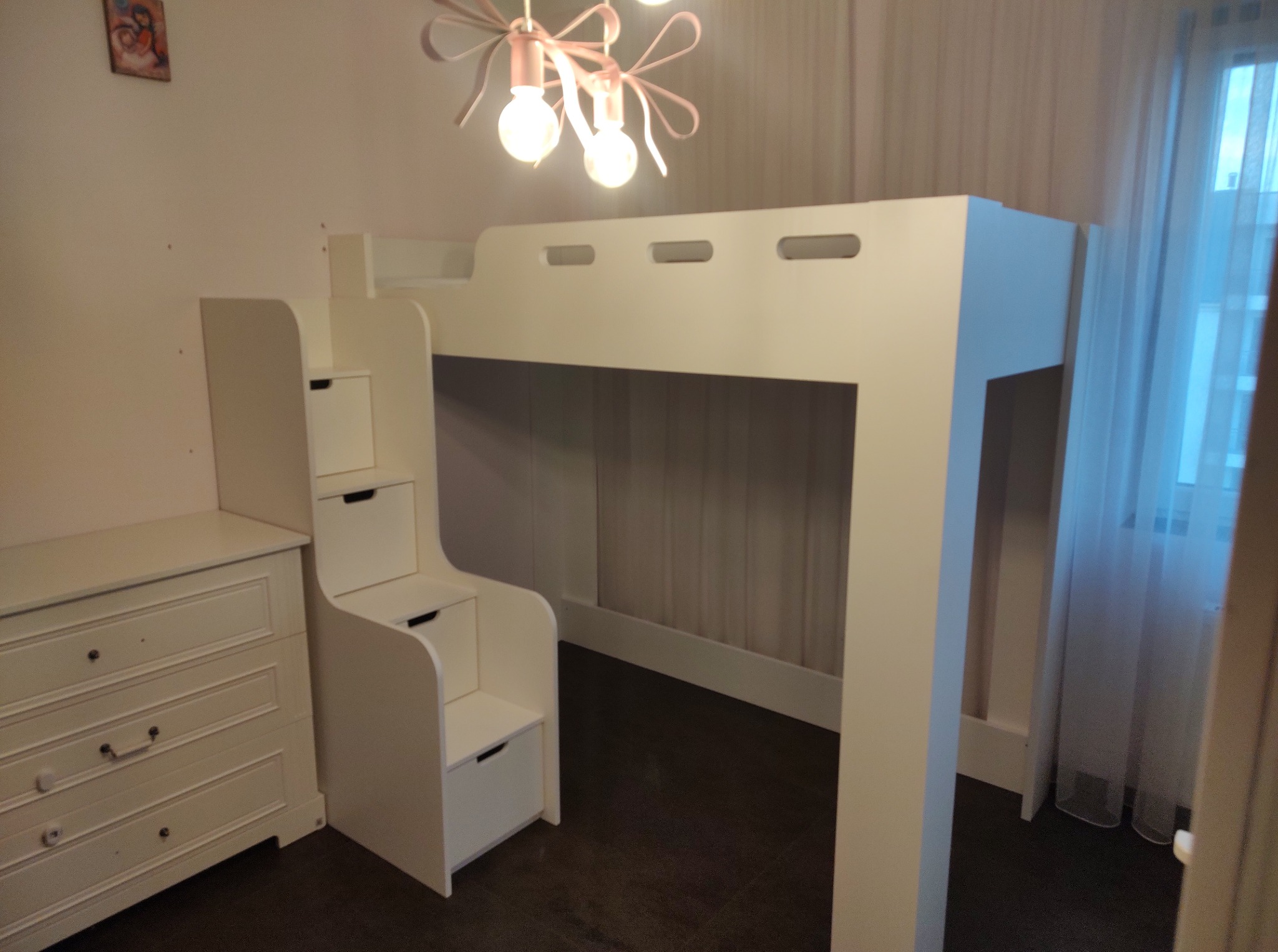 Białe łóżko piętrowe dla dziecka z wbudowanymi schodami-szufladami i komodą, pod różową lampą w pokoju z zasłoniętym oknem.