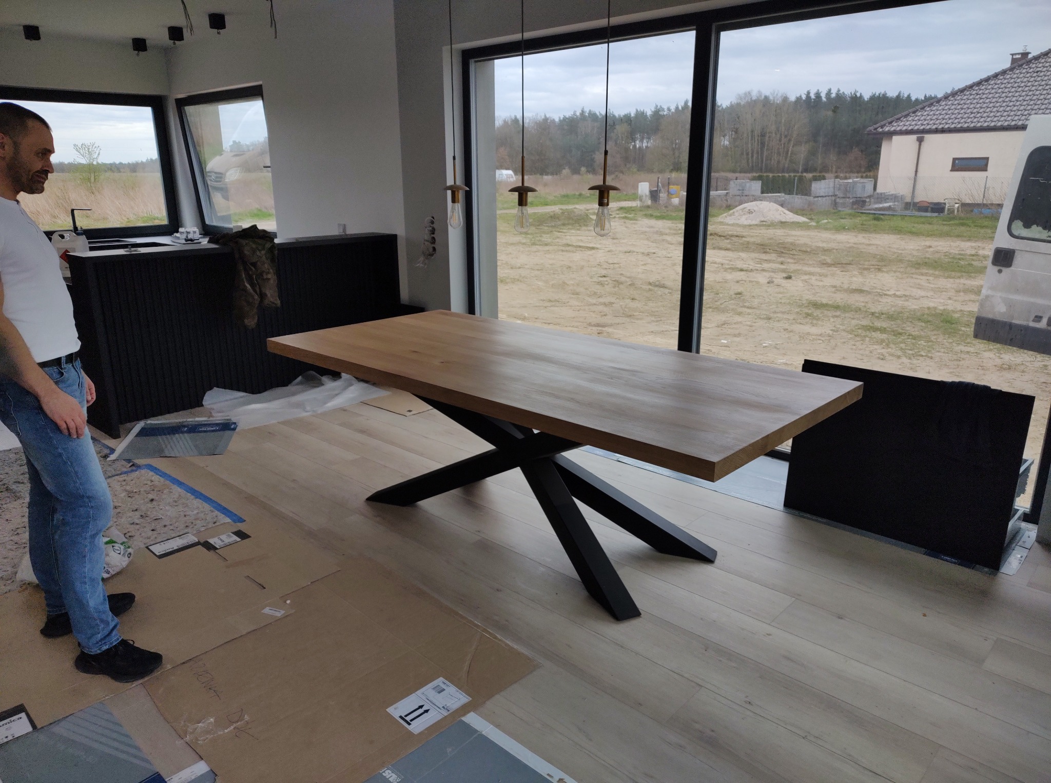 Stół z drewnianym blatem i czarną, geometryczną podstawą w nowoczesnym wnętrzu z dużymi oknami. Widoczny mężczyzna w jeansach i biała koszulka.