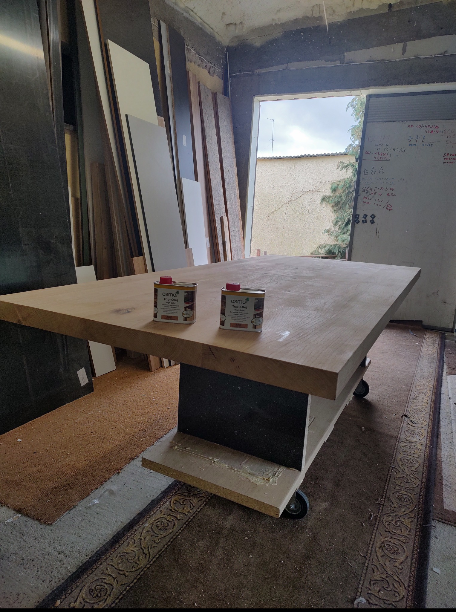 Stolarski warsztat: blat stołu na kółkach z dwiema puszkami oleju Osmo Top-Olej, w tle deski, drzwi i tablica z notatkami, ukazujące etap wykończenia mebla.