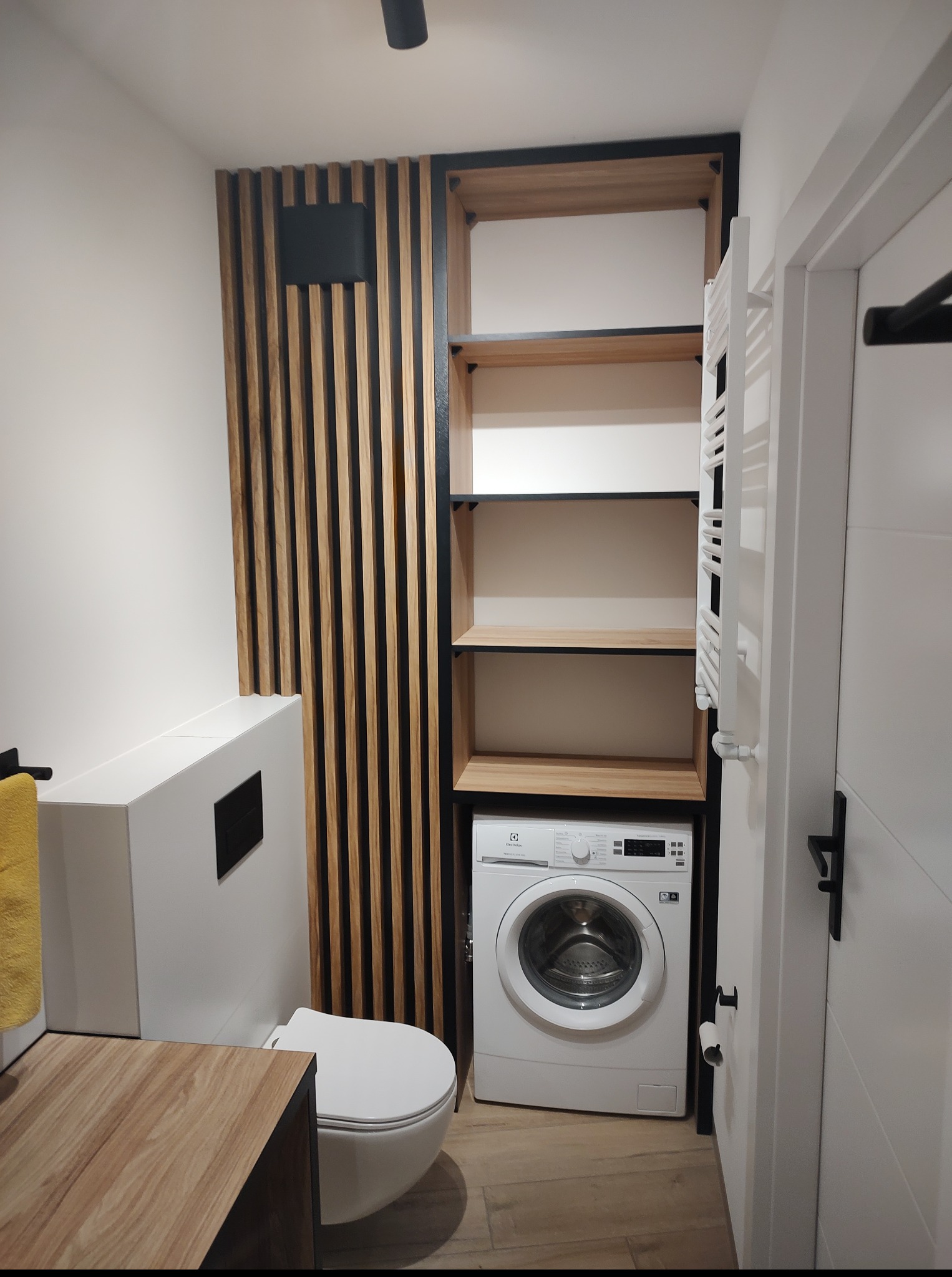 Nowoczesna łazienka z drewnianymi lamelami i półkami na wymiar, z pralką w zabudowie. Minimalistyczny design, czarne akcenty, biała armatura.