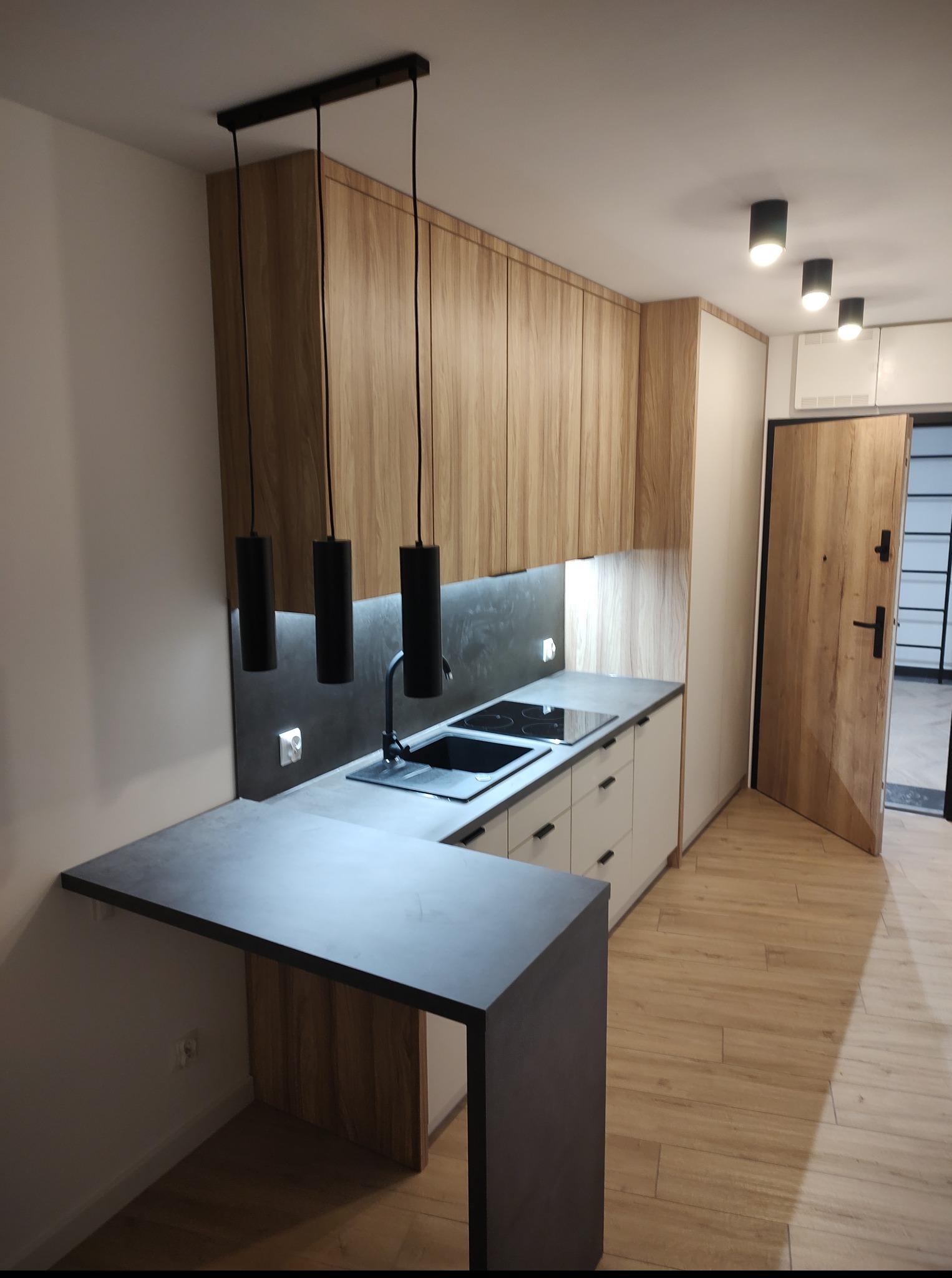 Nowoczesna kuchnia z drewnianymi szafkami wiszącymi, czarnym blatem i minimalistycznym oświetleniem. Widoczny fragment wyspy kuchennej i drewniana podłoga.