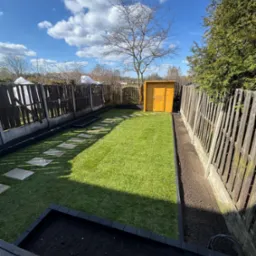 Uporządkowany ogród z równo przystrzyżonym trawnikiem, ścieżką z płyt, drewnianym domkiem narzędziowym i rabatami wzdłuż płotu, w słoneczny dzień.