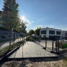 Ułożona kostka brukowa w nowoczesnym stylu, widok z góry na posesję z nowymi domami. Słońce odbija się od kostki. Widoczny fragment trawnika i ogrodzenie.