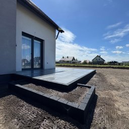 HEKTAR - Nowoczesny taras z ciemnej kostki brukowej, schody z obrzeżami, przygotowany teren pod trawnik, dom jednorodzinny w tle, słoneczny dzień.