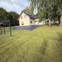 HEKTAR - Nowo założony trawnik przed domem jednorodzinnym, z widocznymi drzewami i czarną folią zabezpieczającą skarpę. Świeża zieleń trawy kontrastuje z jasną elewacją budynku.